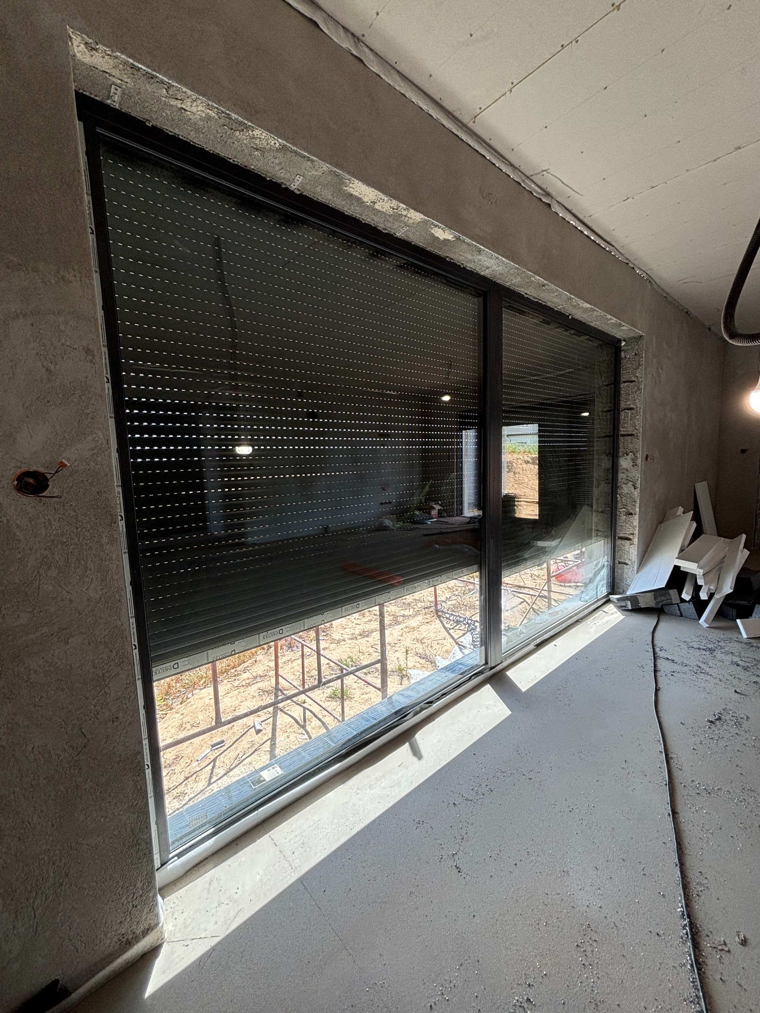 Duże okno przesuwne z czarną ramą i opuszczoną roletą zewnętrzną w surowym wnętrzu. Widoczne niedokończone ściany i podłoga w trakcie prac wykończeniowych.