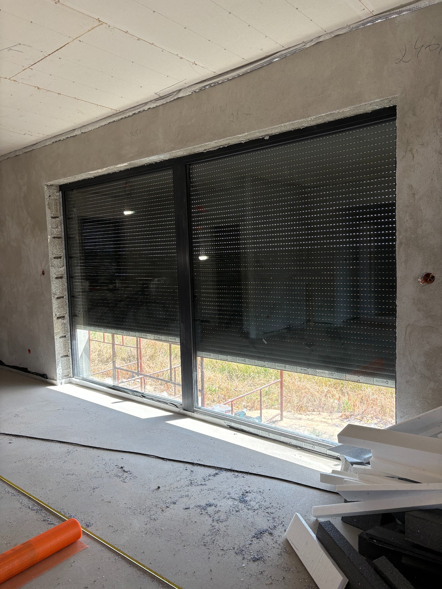 Zamknięta roleta zewnętrzna w oknie z czarną ramą, widok z wnętrza surowego pomieszczenia w trakcie wykończenia. Na podłodze resztki materiałów budowlanych.