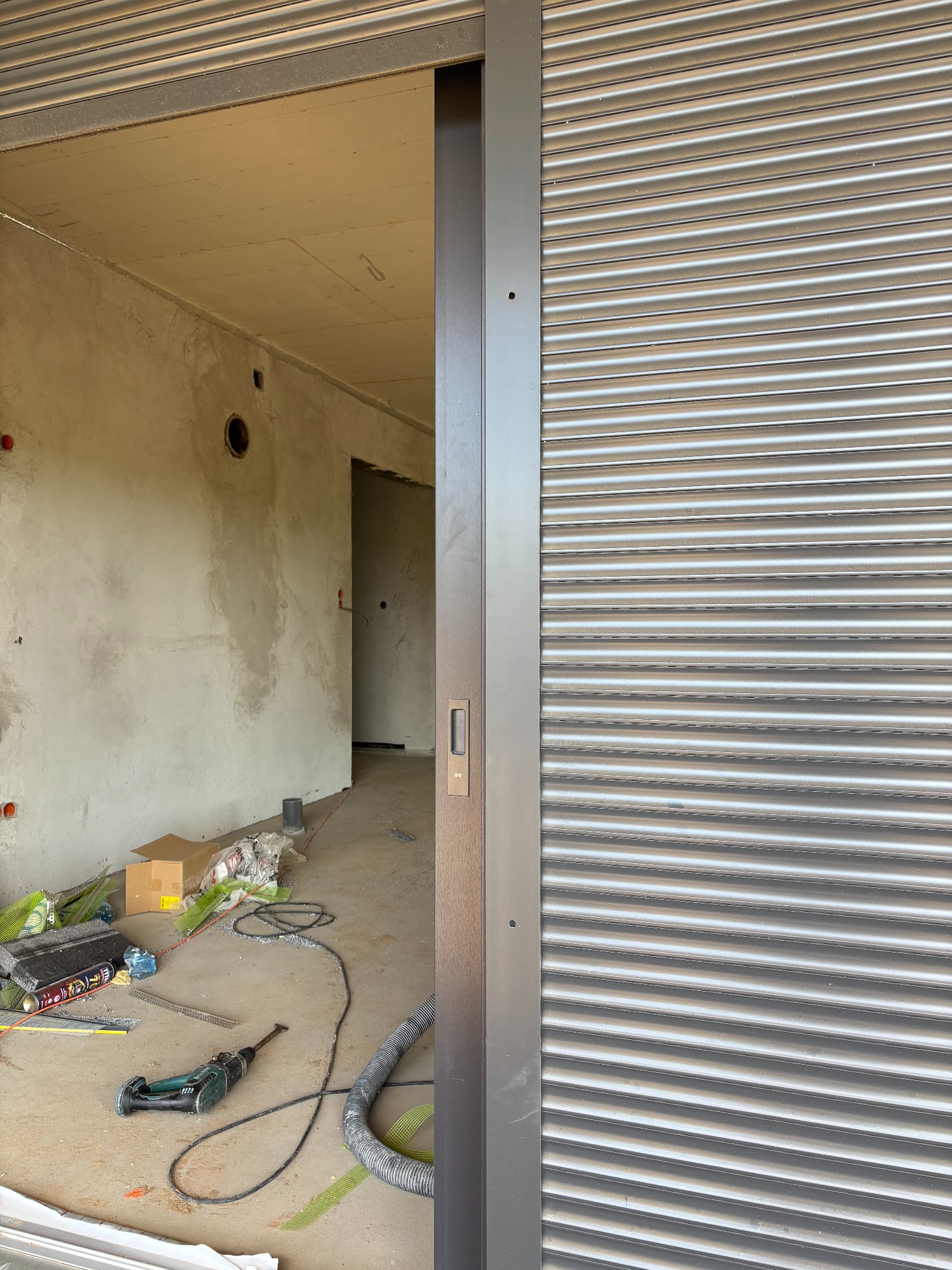 Zamknięta, szara roleta wewnętrzna w surowym wnętrzu. Widoczne narzędzia i materiały budowlane na podłodze. W tle ściany w stanie deweloperskim.