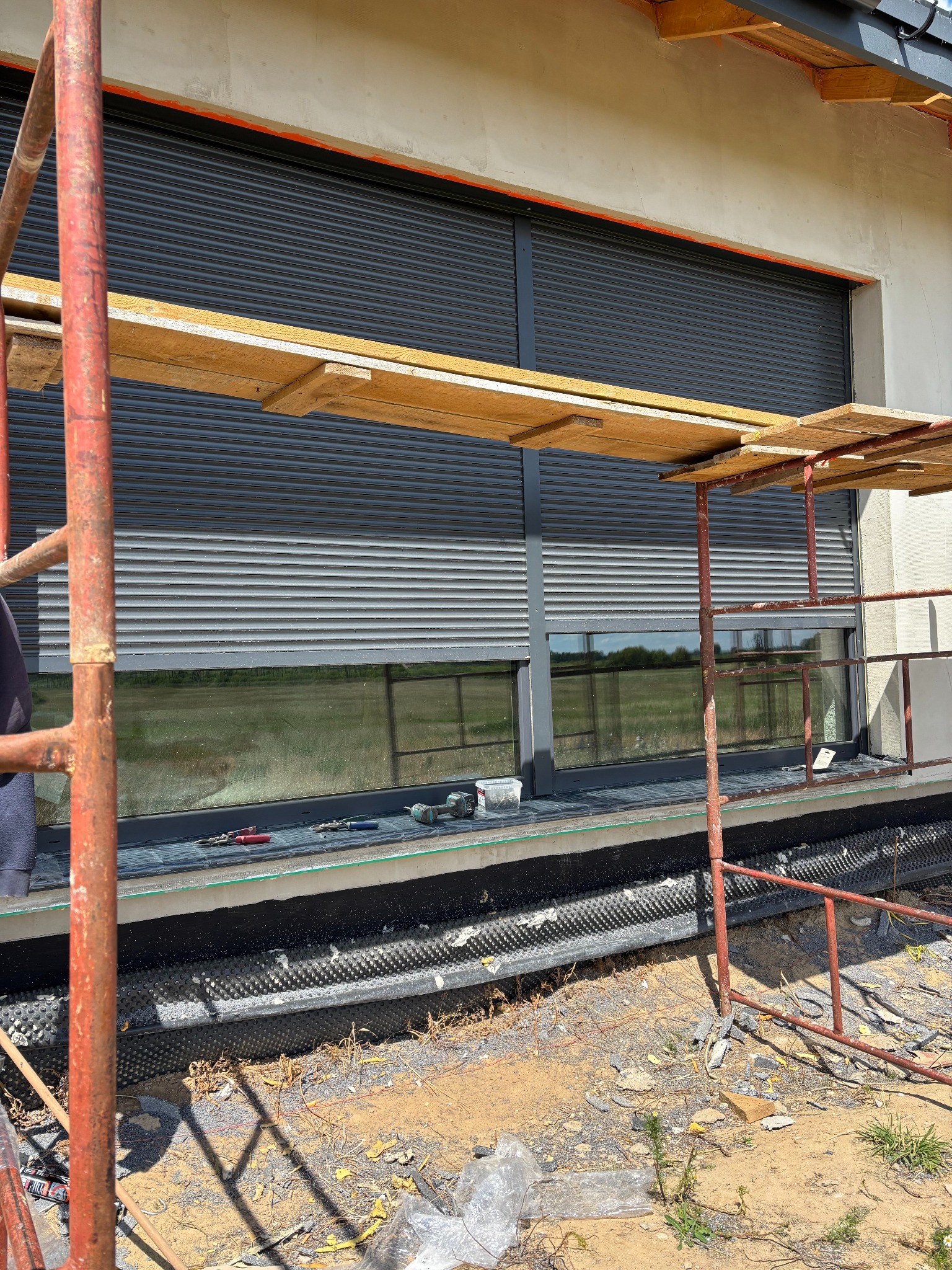 Montaż szarej rolety zewnętrznej na oknie budynku w trakcie budowy, widoczne rusztowanie i narzędzia. Ujęcie z dołu, w tle zielony krajobraz.