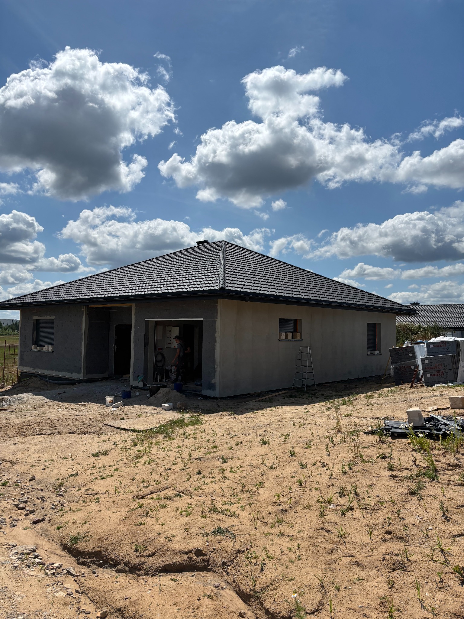 Surowy dom jednorodzinny z ciemną dachówką, widoczne okna z roletami zewnętrznymi, teren wokół w trakcie prac wykończeniowych, błękitne niebo z chmurami.