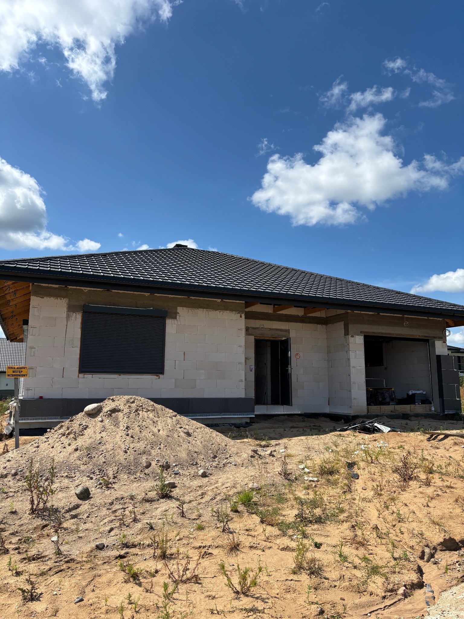 Dom w trakcie budowy z zamontowaną roletą zewnętrzną. Widoczny surowy stan ścian z betonu komórkowego, czarna dachówka i sterta piasku na pierwszym planie.