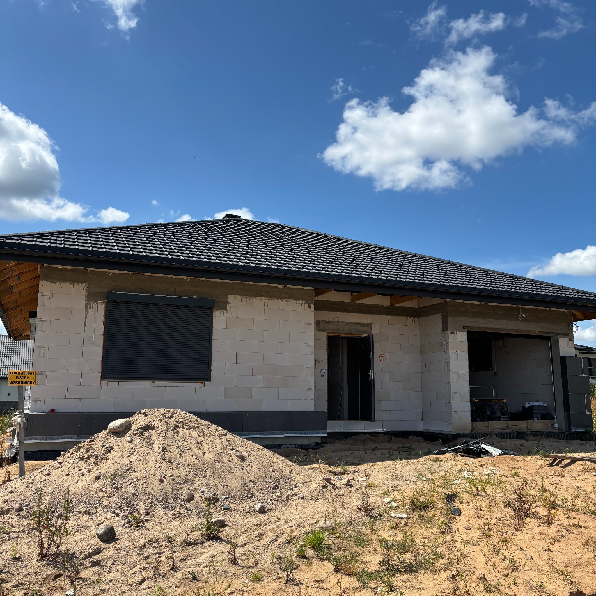 Dom w trakcie budowy z szarej kostki, z zamontowaną roletą zewnętrzną. Na pierwszym planie hałda piasku. Błękitne niebo z chmurami w tle.