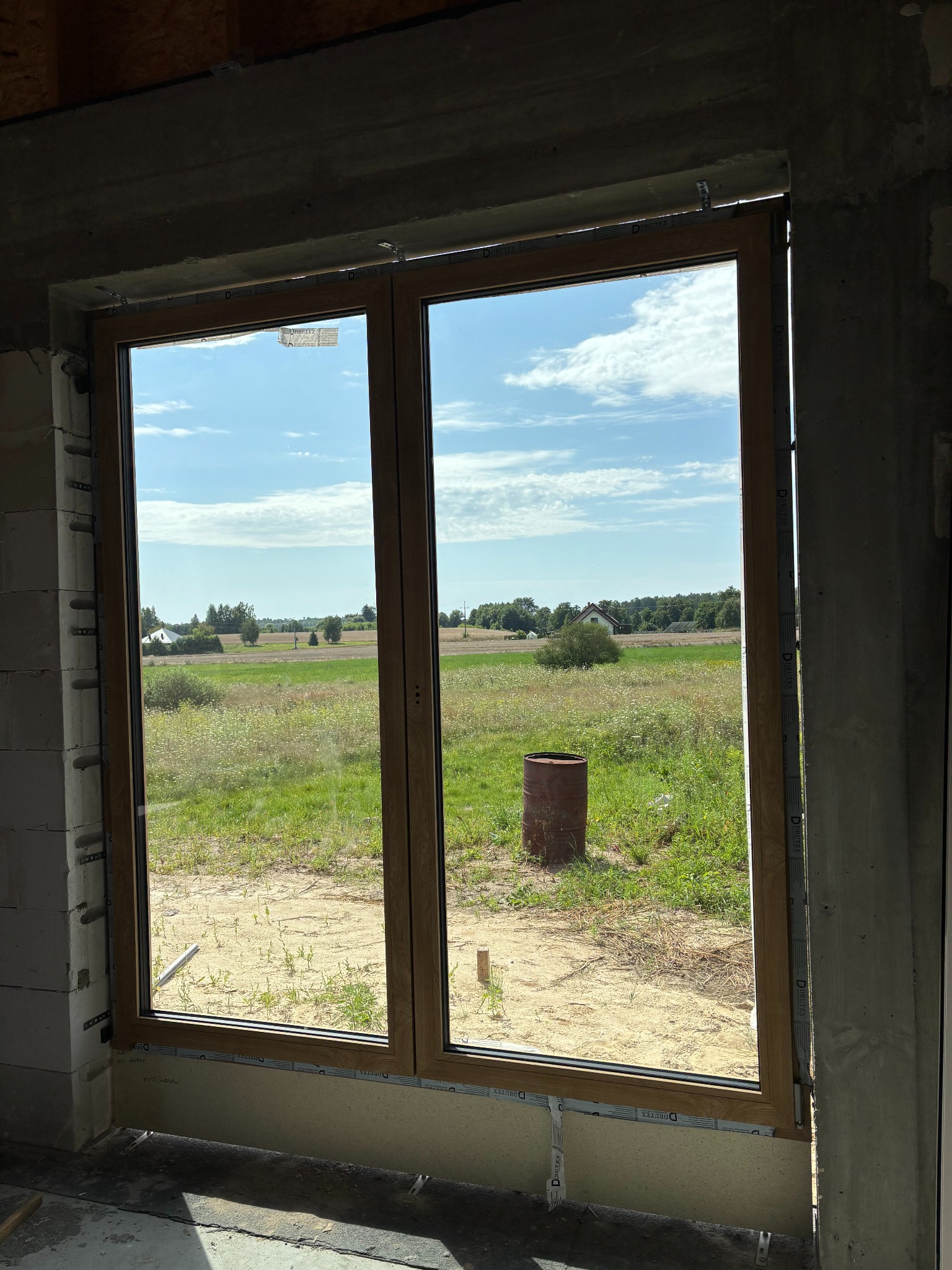 Zamontowane okno dwuskrzydłowe z drewnianą ramą w surowym otworze budynku, widok na łąkę i niebo. Widoczne detale montażowe.
