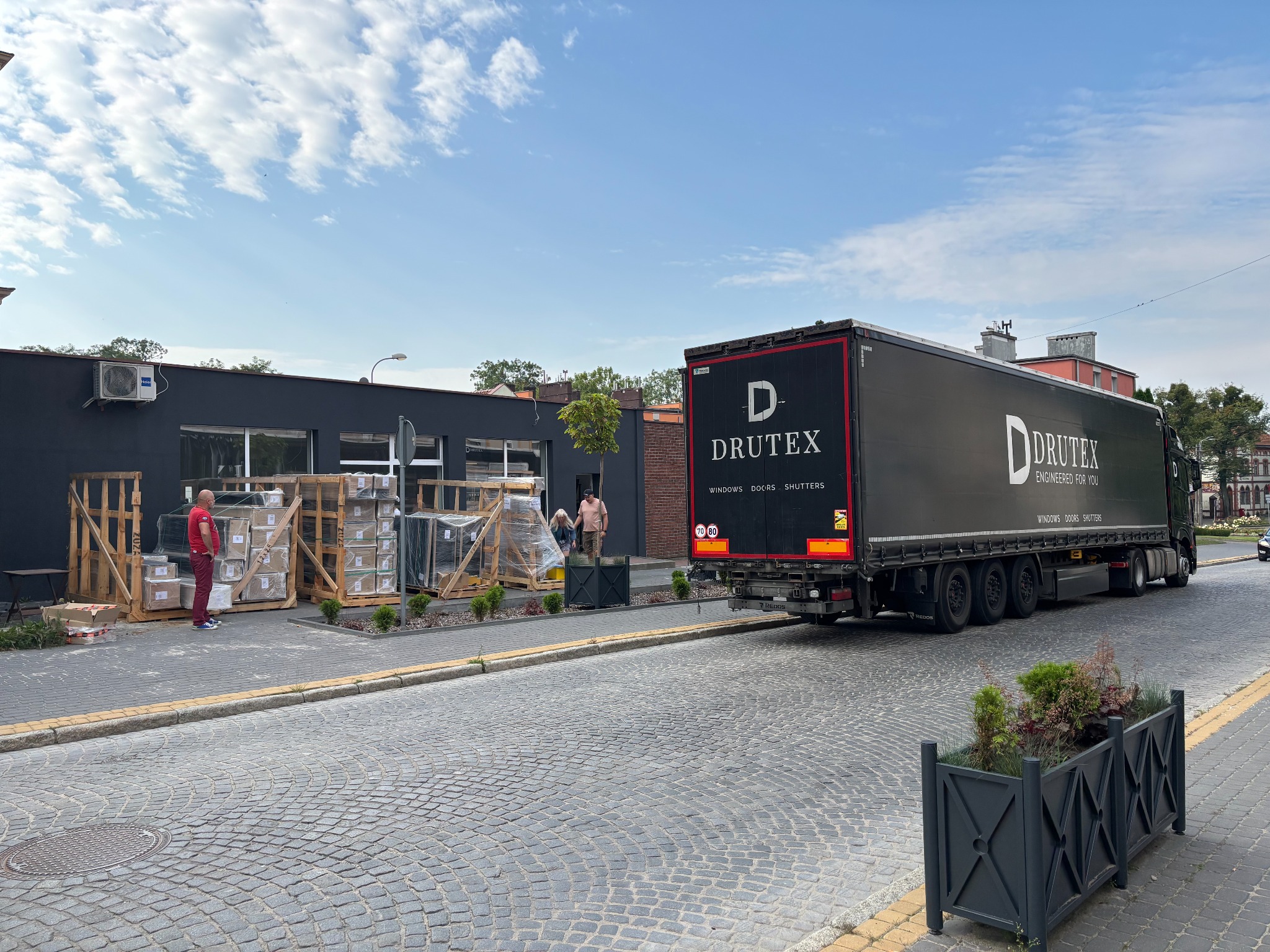 Ciężarówka Drutex z oknami i drzwiami na brukowanej ulicy. Palety z oknami czekają na rozładunek przed budynkiem. Widoczni ludzie i elementy otoczenia.