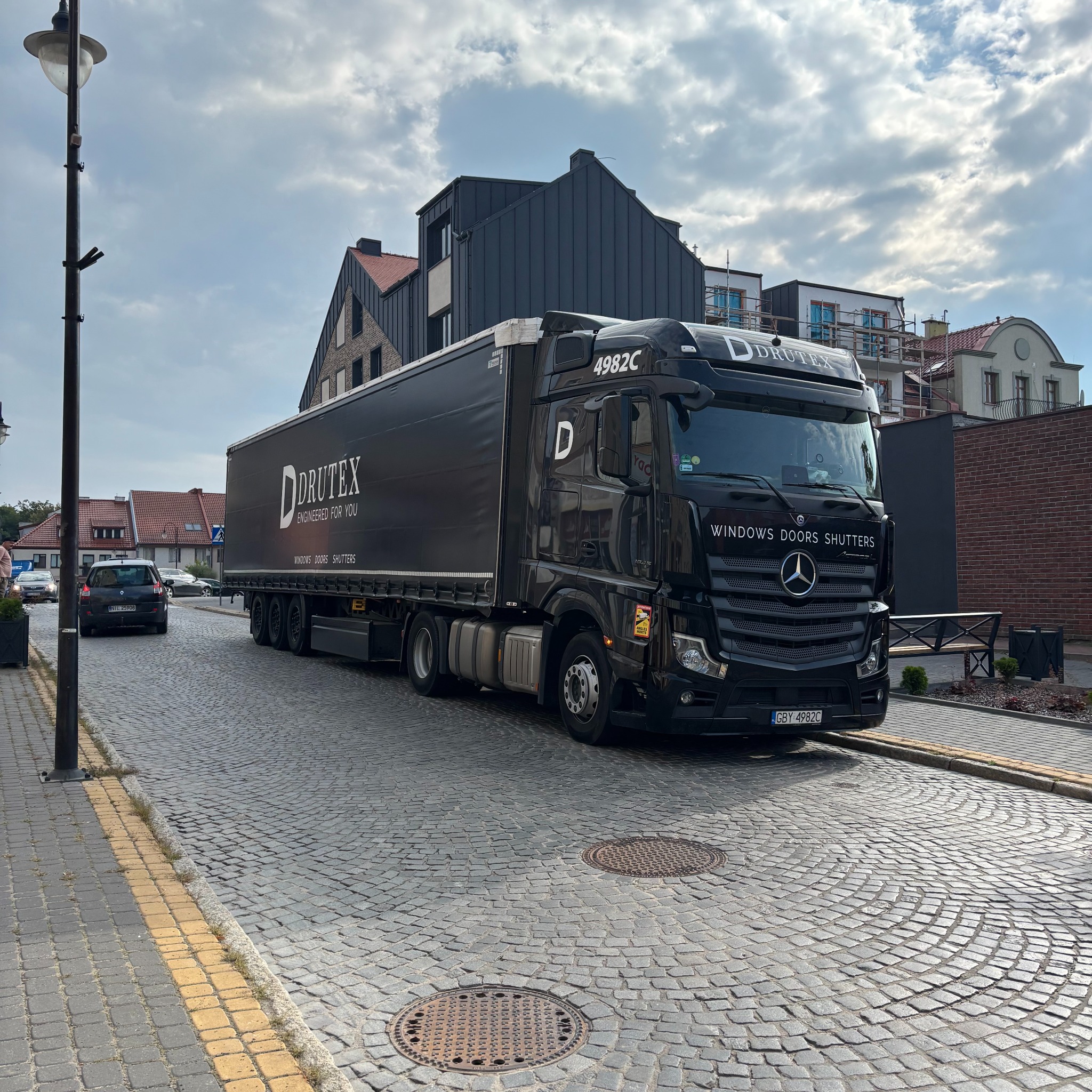 Czarna ciężarówka z logo Drutex na tle miejskiej ulicy z kostki brukowej i budynków. Widoczne napisy: Windows, Doors, Shutters.