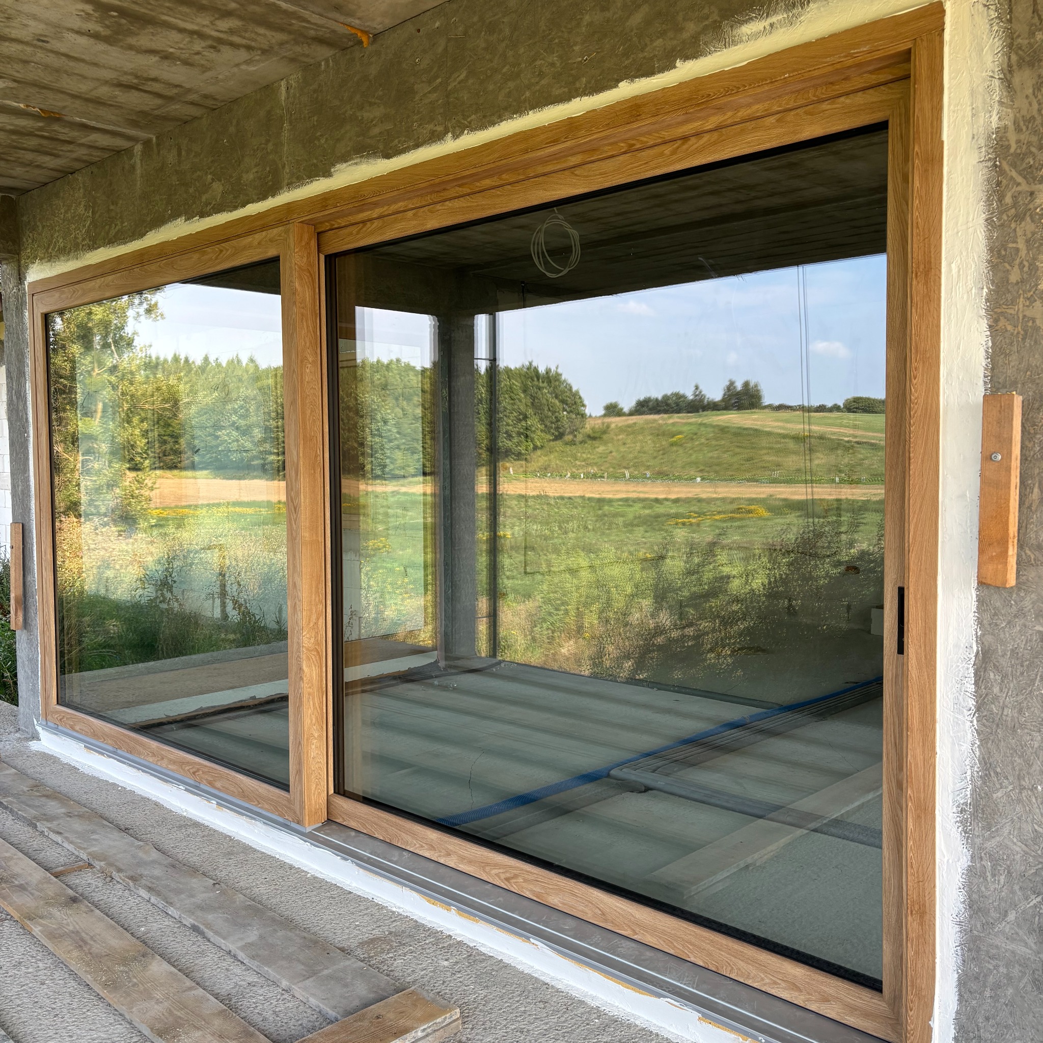 Duże okno przesuwne z drewnianą ramą, widok na zielony krajobraz. Surowy stan wykończenia wnętrza i elewacji. Widoczne elementy instalacji.