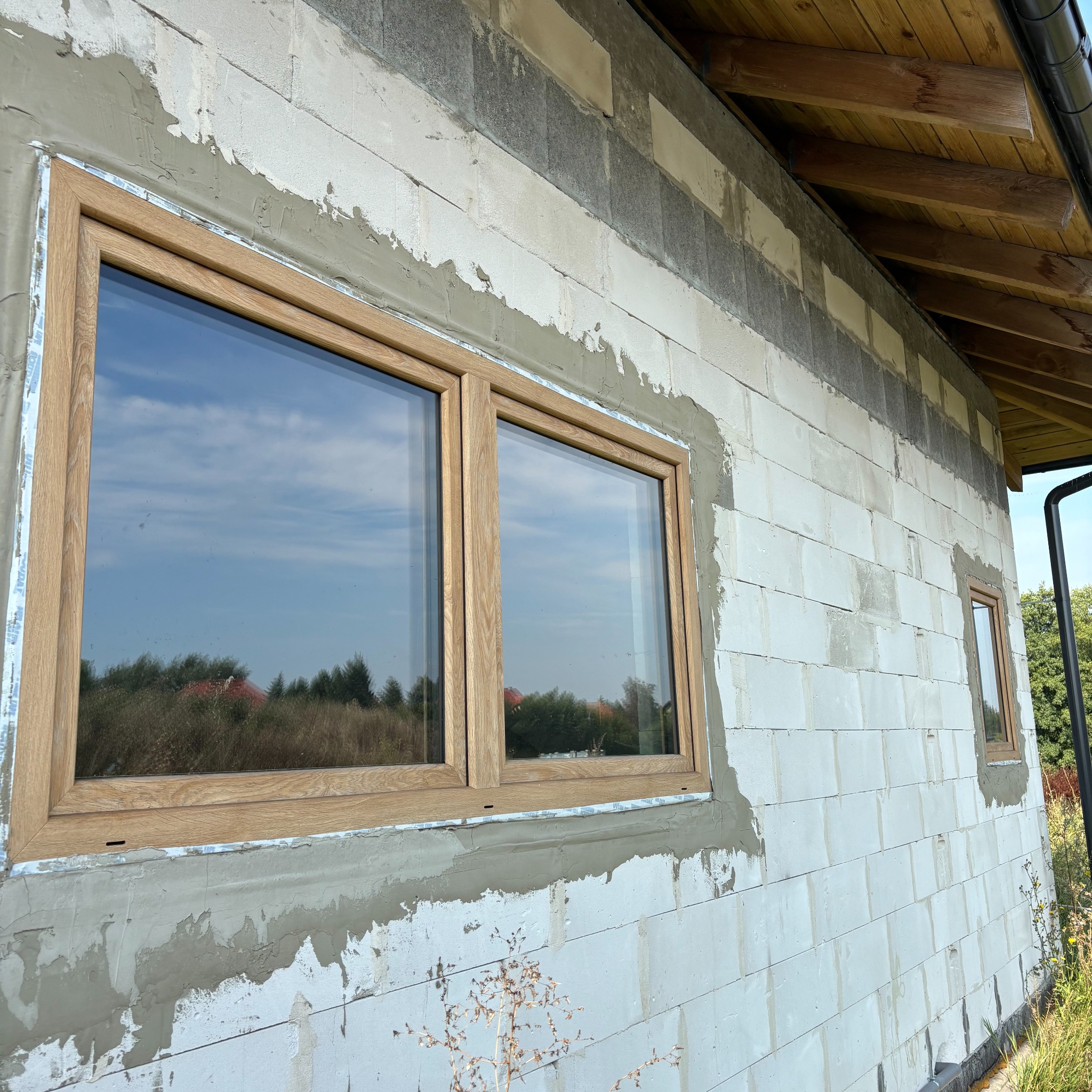 Dwa okna z drewnianą ramą w surowej ścianie z betonu komórkowego, widoczne ślady po montażu. W szybach odbija się niebo i drzewa.