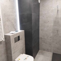 Kacper Czop - Nowoczesna łazienka z szarymi płytkami imitującymi kamień, podwieszana toaleta i czarny, wąski brodzik. Minimalistyczny design z oświetleniem LED.