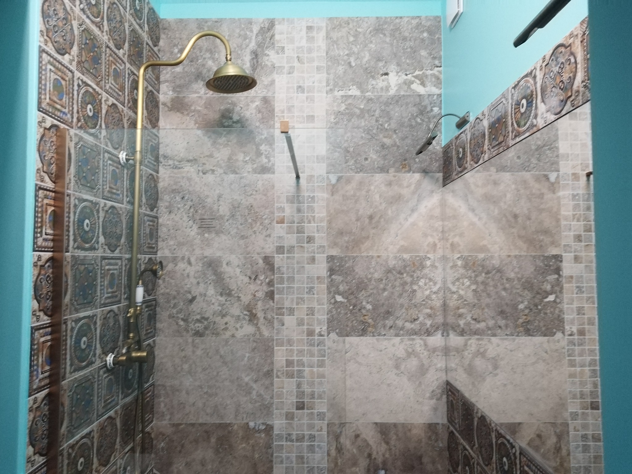 Wykończona kabina prysznicowa z mozaiką i złotą armaturą. Ściany wyłożone płytkami w odcieniach szarości i beżu. Nietypowe wzornictwo płytek dekoracyjnych.