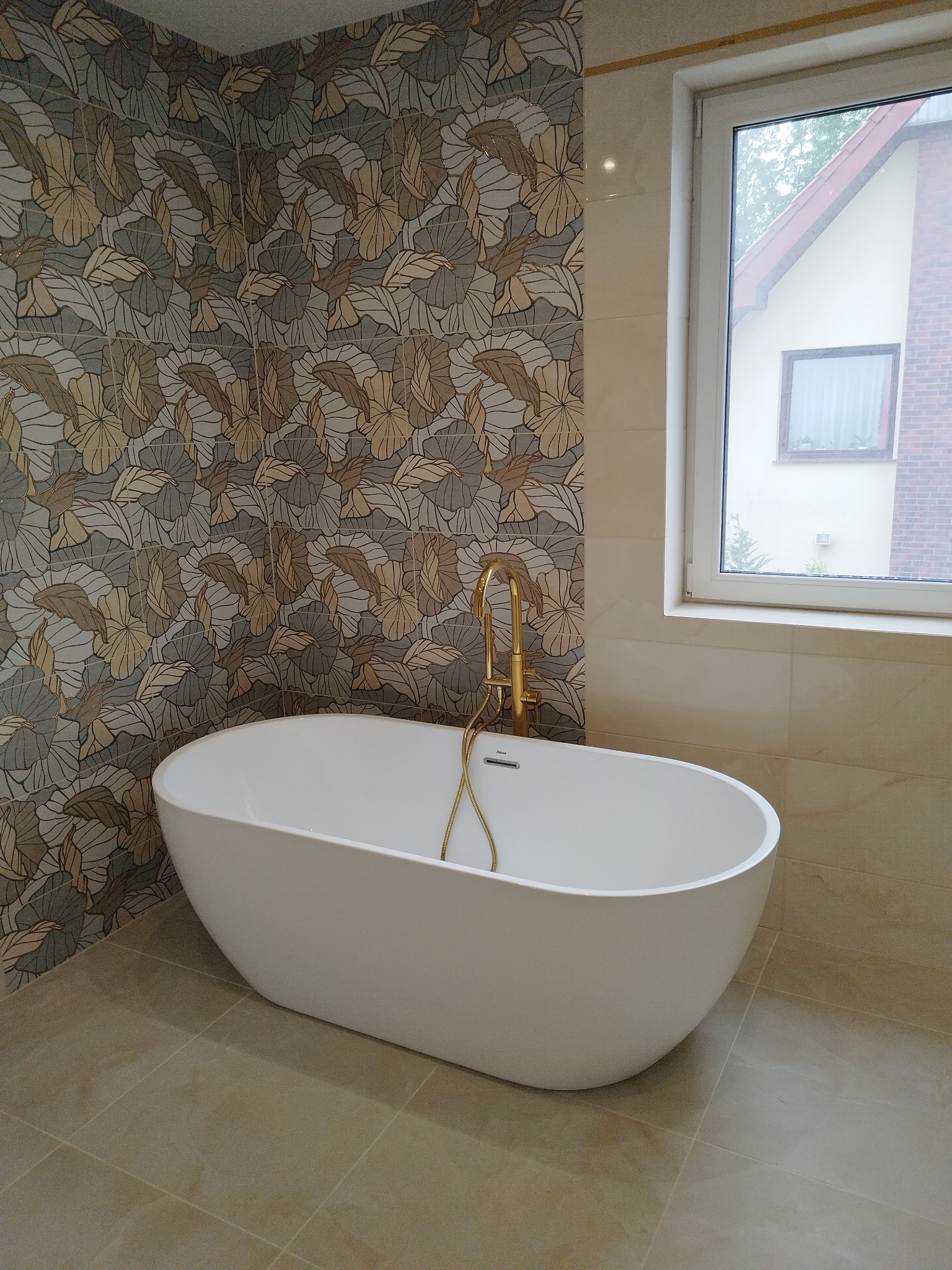Nowoczesna, wolnostojąca wanna owalna z złotą armaturą na tle ściany z dekoracyjnymi płytkami w łazience z oknem. Beżowe płytki podłogowe.