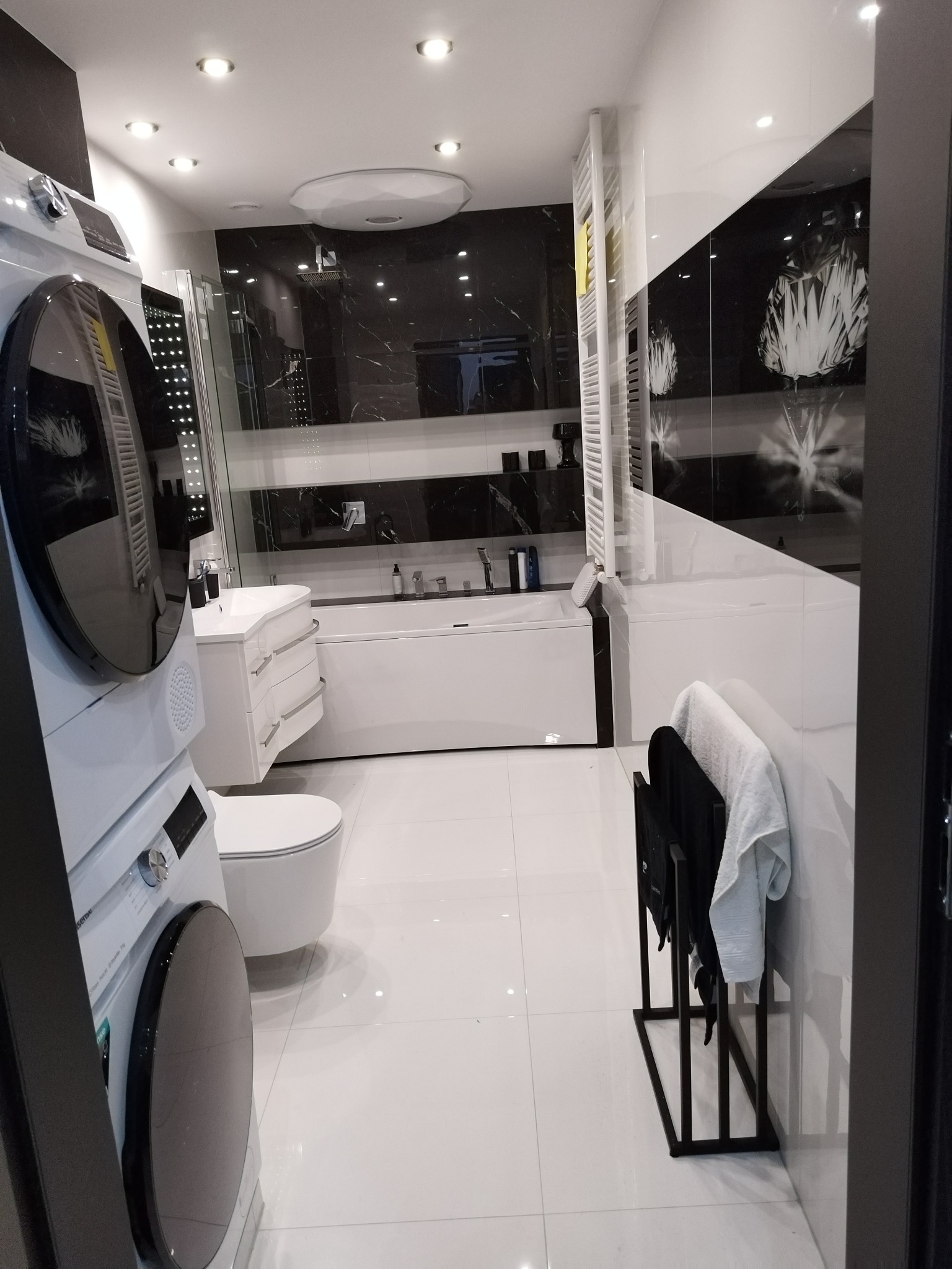 Nowoczesna łazienka w bieli i czerni z pralką na pierwszym planie. Minimalistyczny design, oświetlenie punktowe, wanna i kabina prysznicowa w tle.