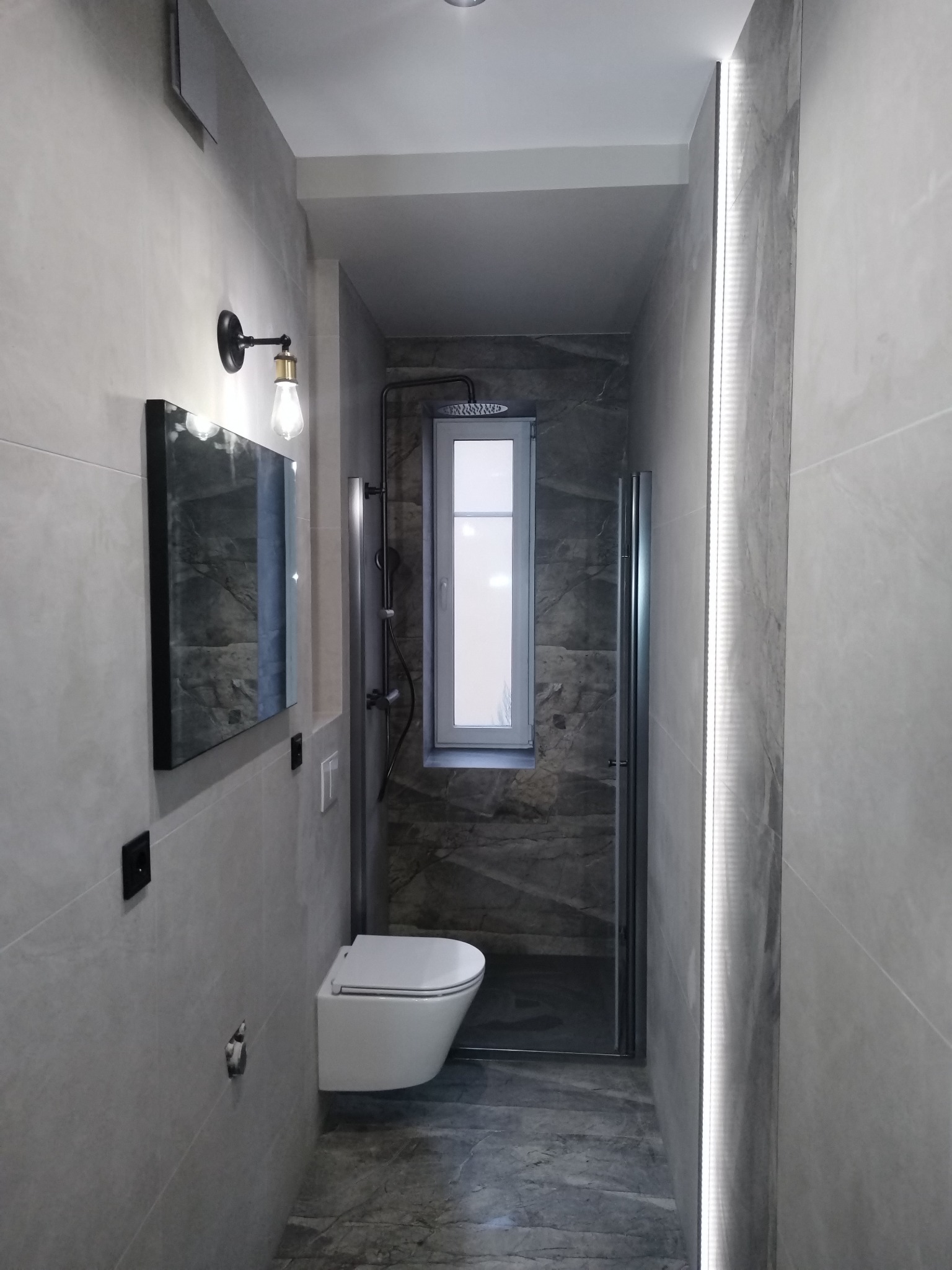 Nowoczesna łazienka w szarościach: podświetlane lustro, wisząca toaleta, kabina prysznicowa z oknem. Minimalistyczny design z akcentami retro w oświetleniu.