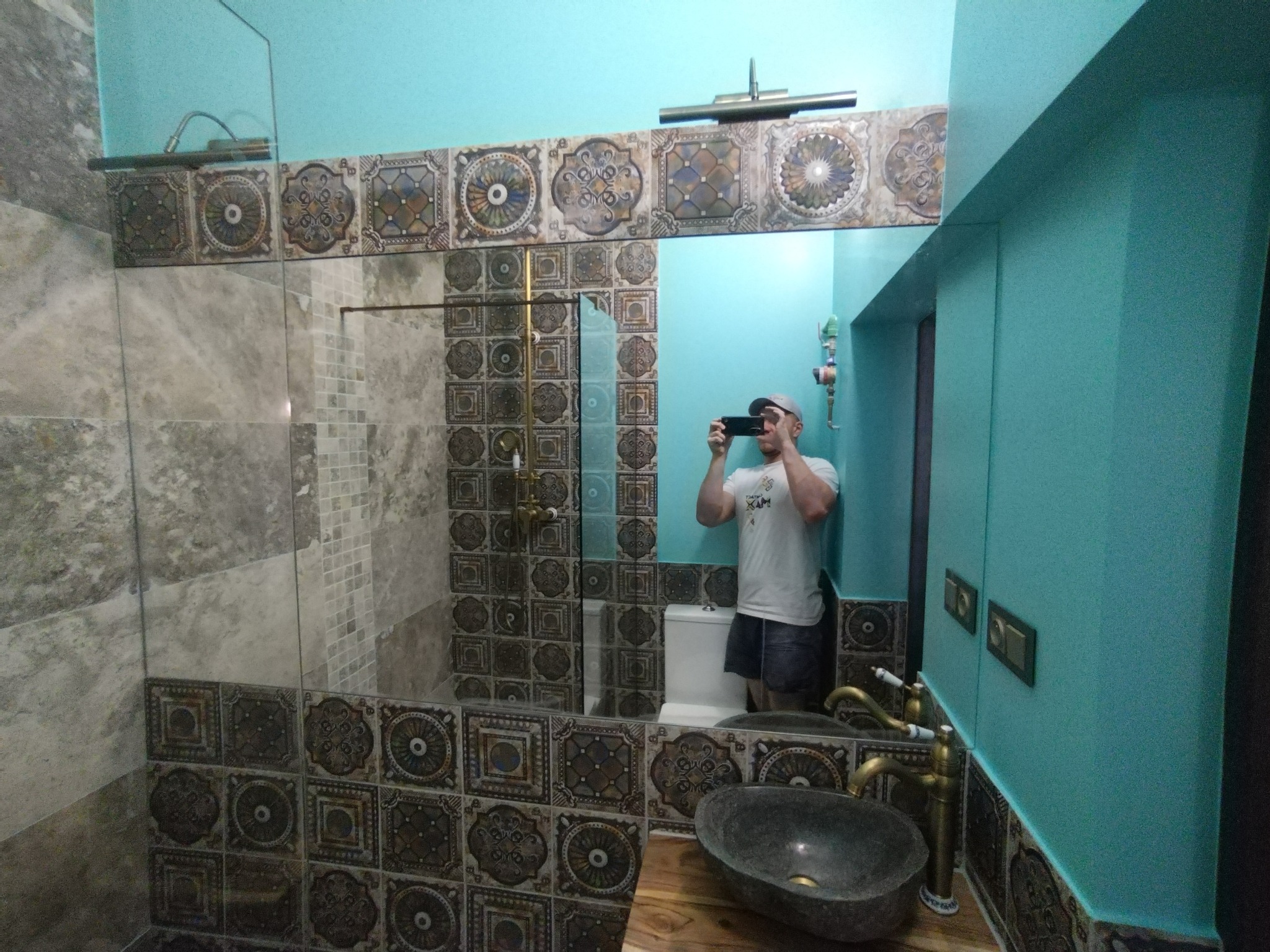 Wykończona łazienka z turkusowymi ścianami i dekoracyjnymi płytkami. Umywalka z kamienia i bateria w stylu retro. W lustrze odbija się osoba robiąca zdjęcie.