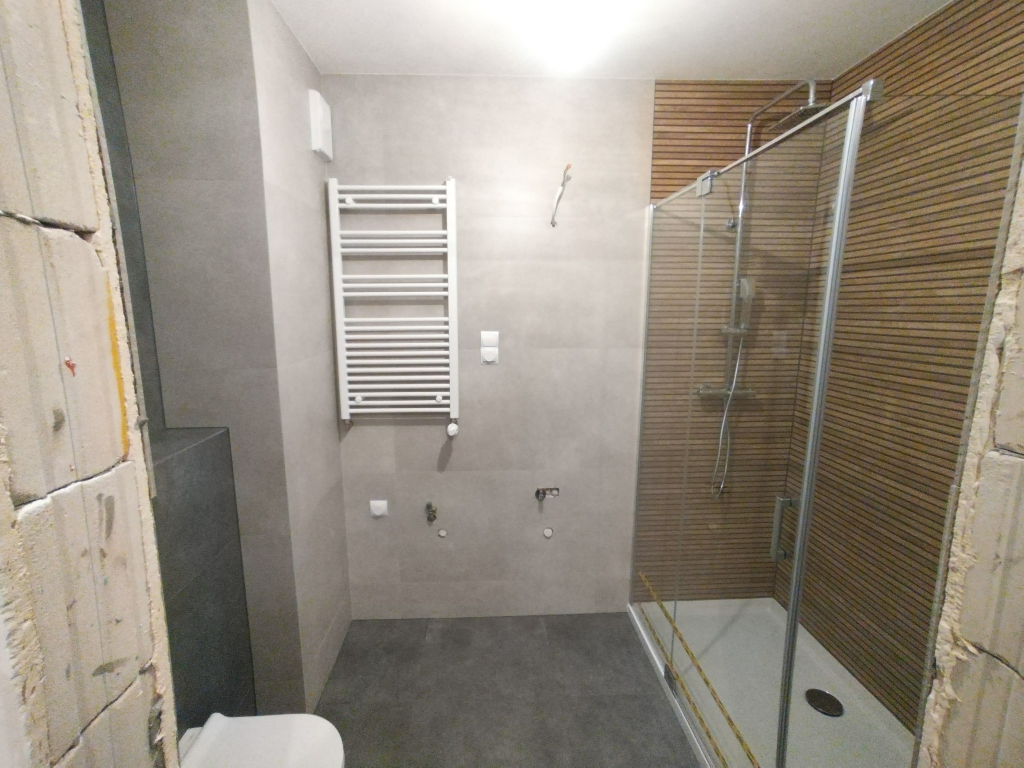 Nowoczesna łazienka w trakcie wykańczania: szare płytki, drewniane panele pod prysznicem, biały grzejnik drabinkowy. Widoczne surowe ściany w tle.