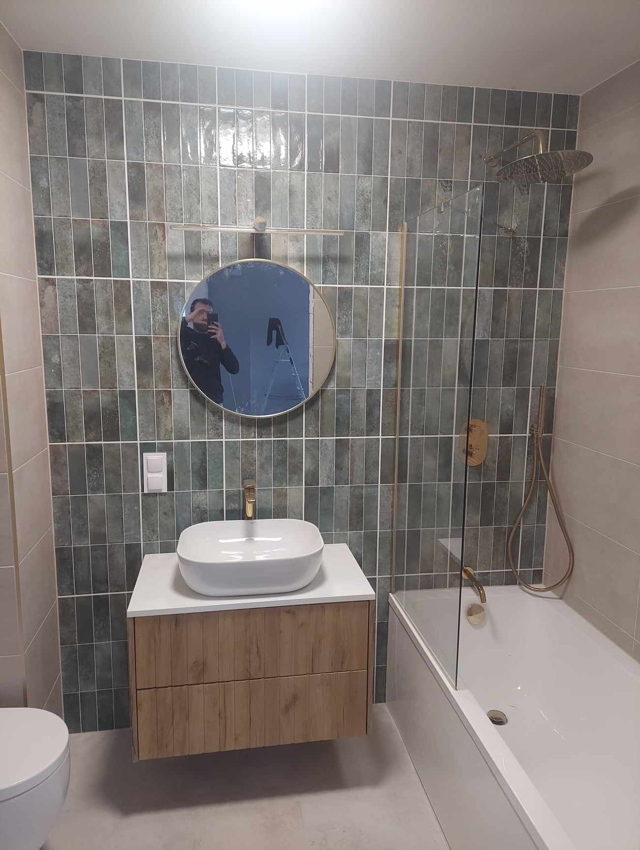 Nowoczesna łazienka z umywalką nablatową, baterią w kolorze złota i zielonymi płytkami ułożonymi pionowo. W lustrze odbija się osoba robiąca zdjęcie.