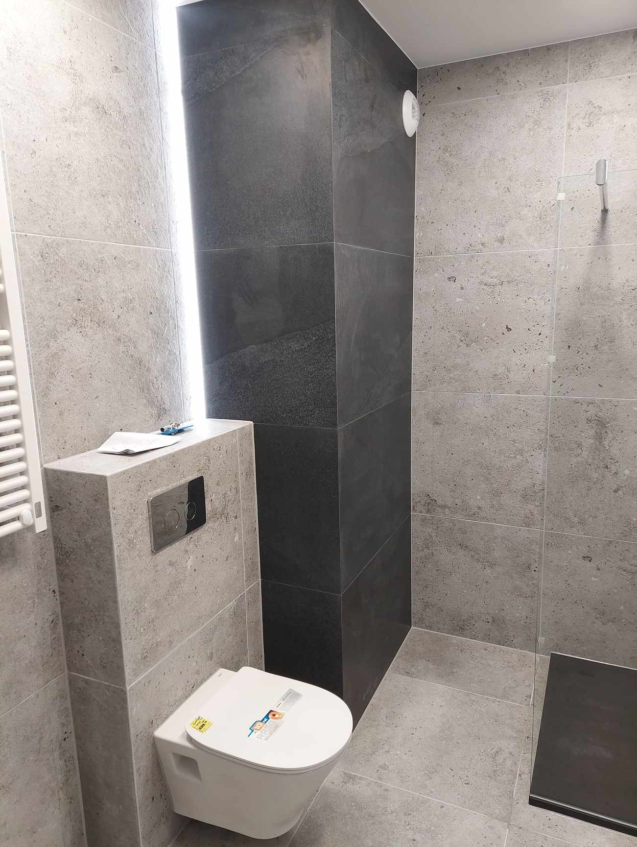 Nowoczesna łazienka z szarymi płytkami imitującymi kamień, podwieszana toaleta i czarny, wąski brodzik. Minimalistyczny design z oświetleniem LED.