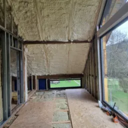 Poddasze w trakcie ocieplania pianką PUR, widoczne krokwie, okno z widokiem na zieleń i narzędzia pracy. Na podłodze płyty OSB i częściowo ułożona wełna.