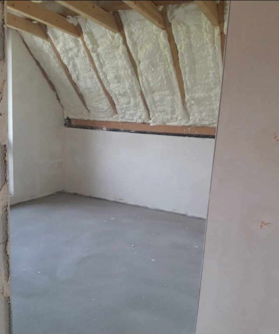 Poddasze po ociepleniu pianką PUR: widoczne drewniane krokwie i biała pianka między nimi. Ściany pomalowane na biało, surowa wylewka podłogowa.