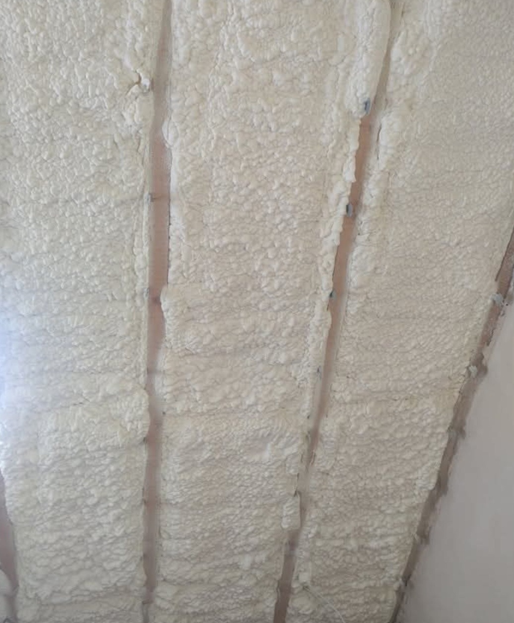 Biała pianka PUR wypełniająca przestrzeń między drewnianymi belkami stropu, widoczna z bliska. Izolacja natryskowa, równomierna struktura.