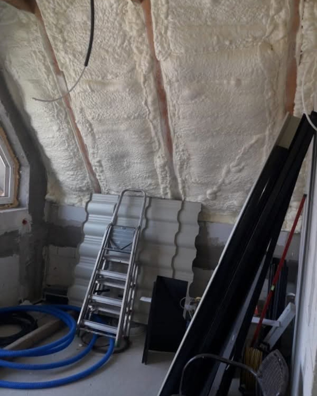 Poddasze po ociepleniu pianką PUR, widoczne równomierne pokrycie powierzchni. Na podłodze drabina i niebieski przewód, okno w tle. Detal wykonanej izolacji.