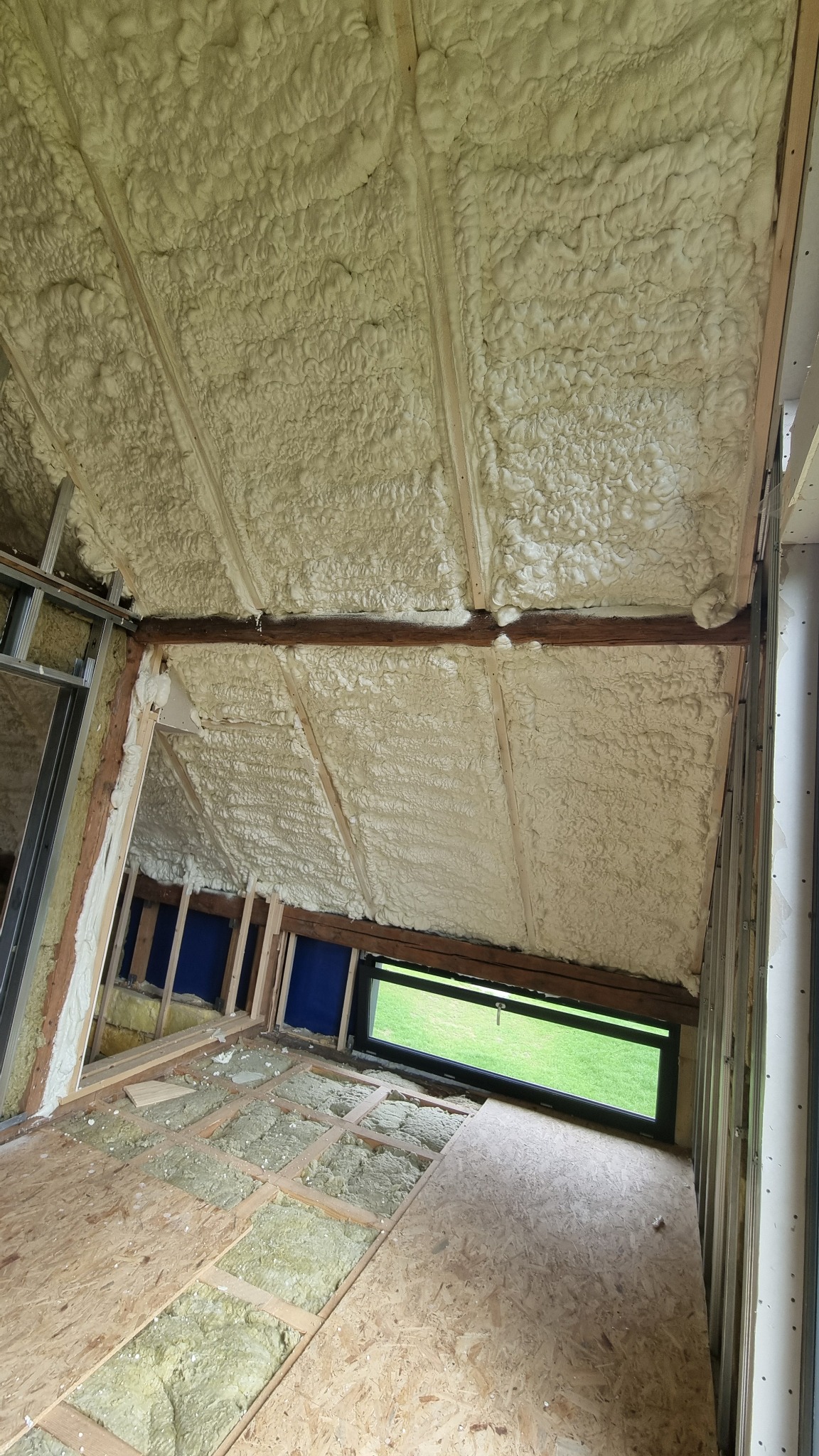 Poddasze z widoczną warstwą izolacji pianką PUR na skośnym dachu, drewnianą konstrukcją i oknem. Częściowo ułożona podłoga z izolacją.