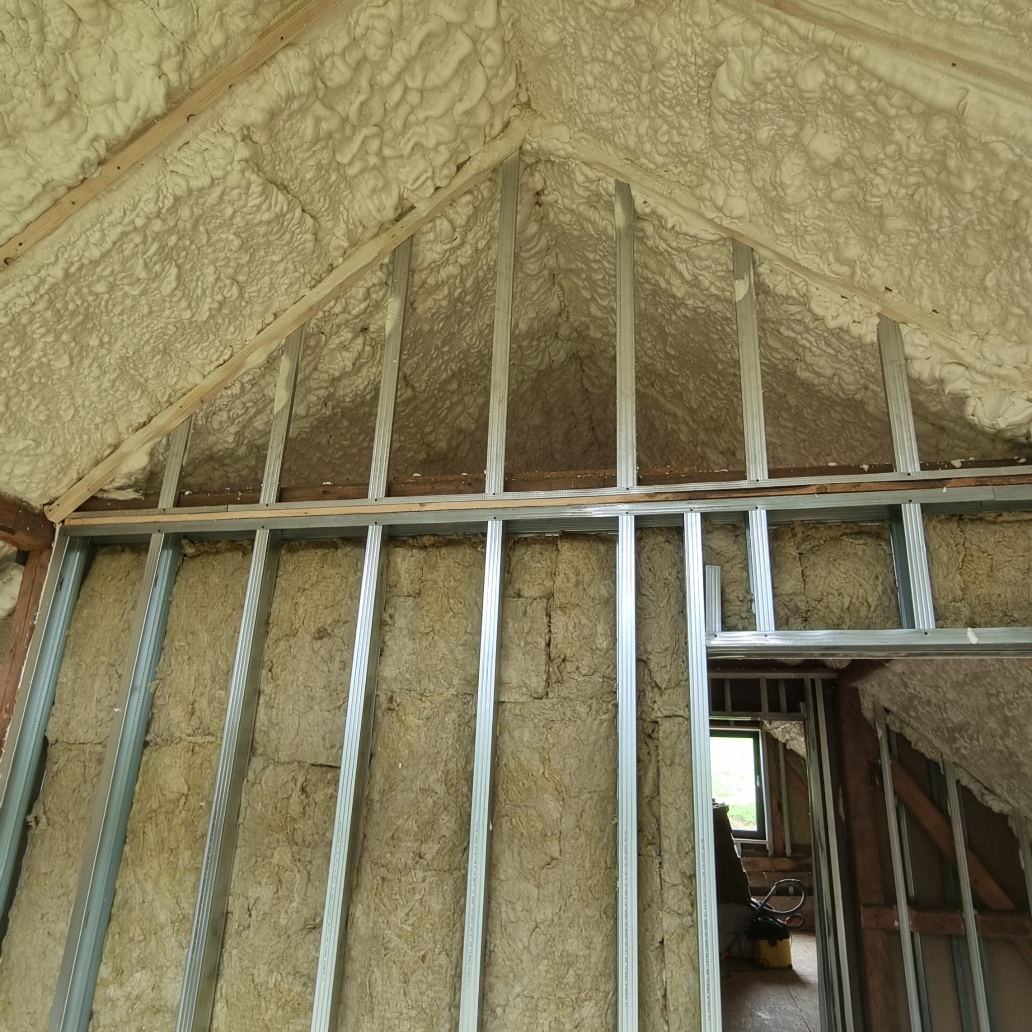 Poddasze z widoczną izolacją: pianka PUR na skosach i wełna mineralna między metalowymi profilami na ścianach. Widoczne okno i elementy konstrukcji.