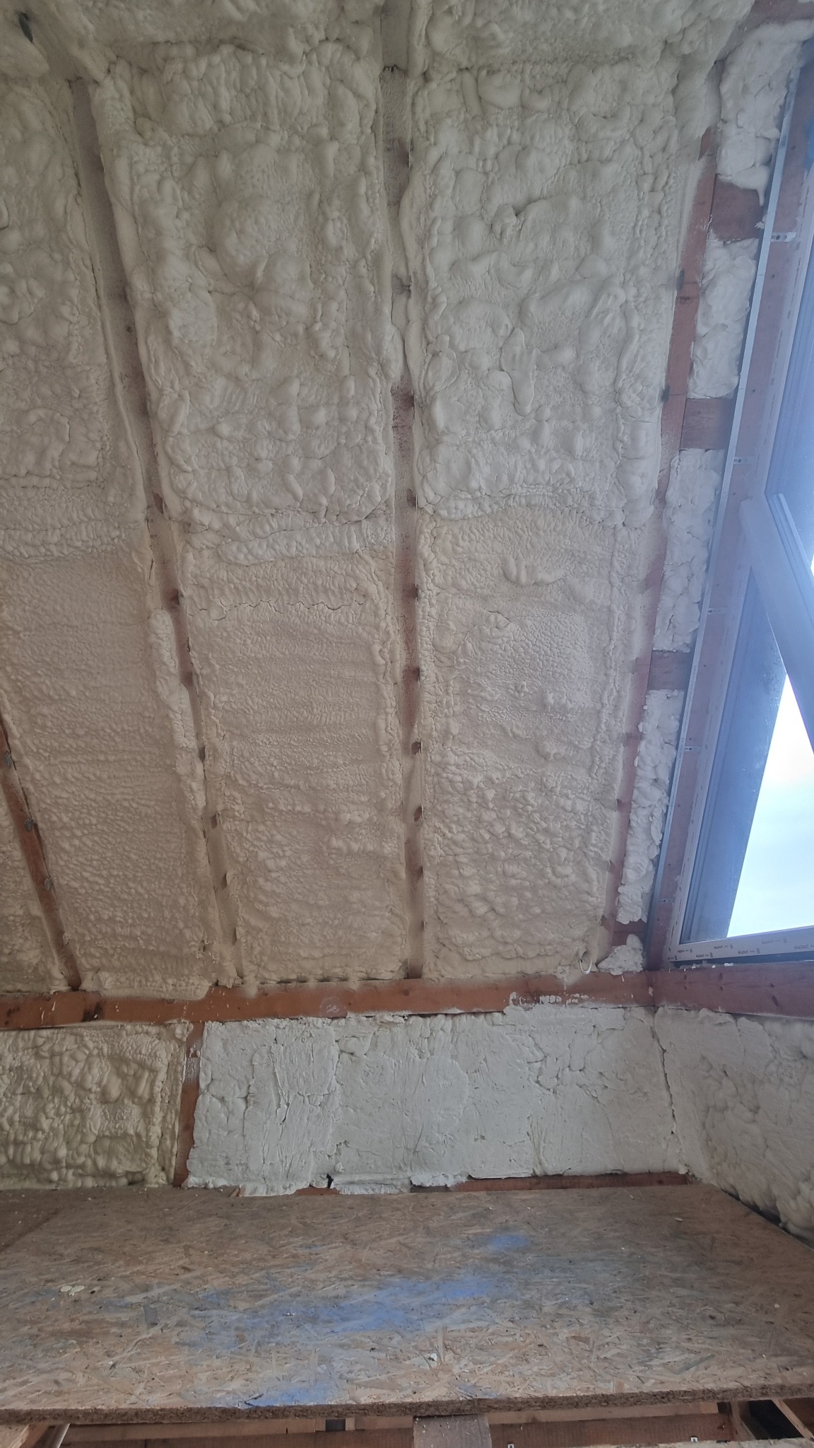 Poddasze po izolacji pianką PUR, widoczne drewniane belki i okno dachowe. Pianka wypełnia przestrzenie między belkami, tworząc jednolitą warstwę izolacyjną.