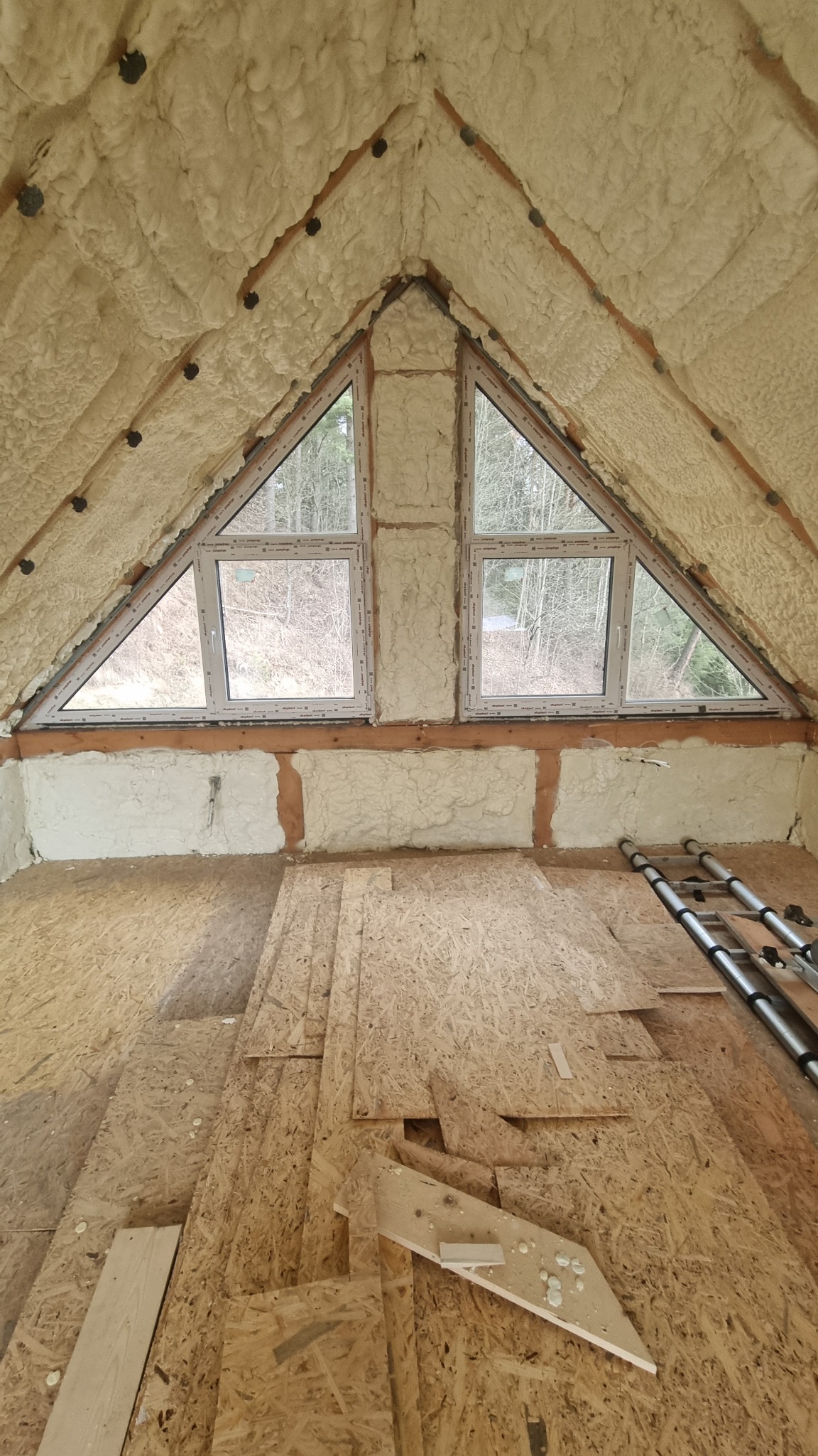 Poddasze z oknami trójkątnymi, ocieplone pianką PUR, widoczne płyty OSB na podłodze i drabina. Izolacja poddasza z nietypowymi oknami.