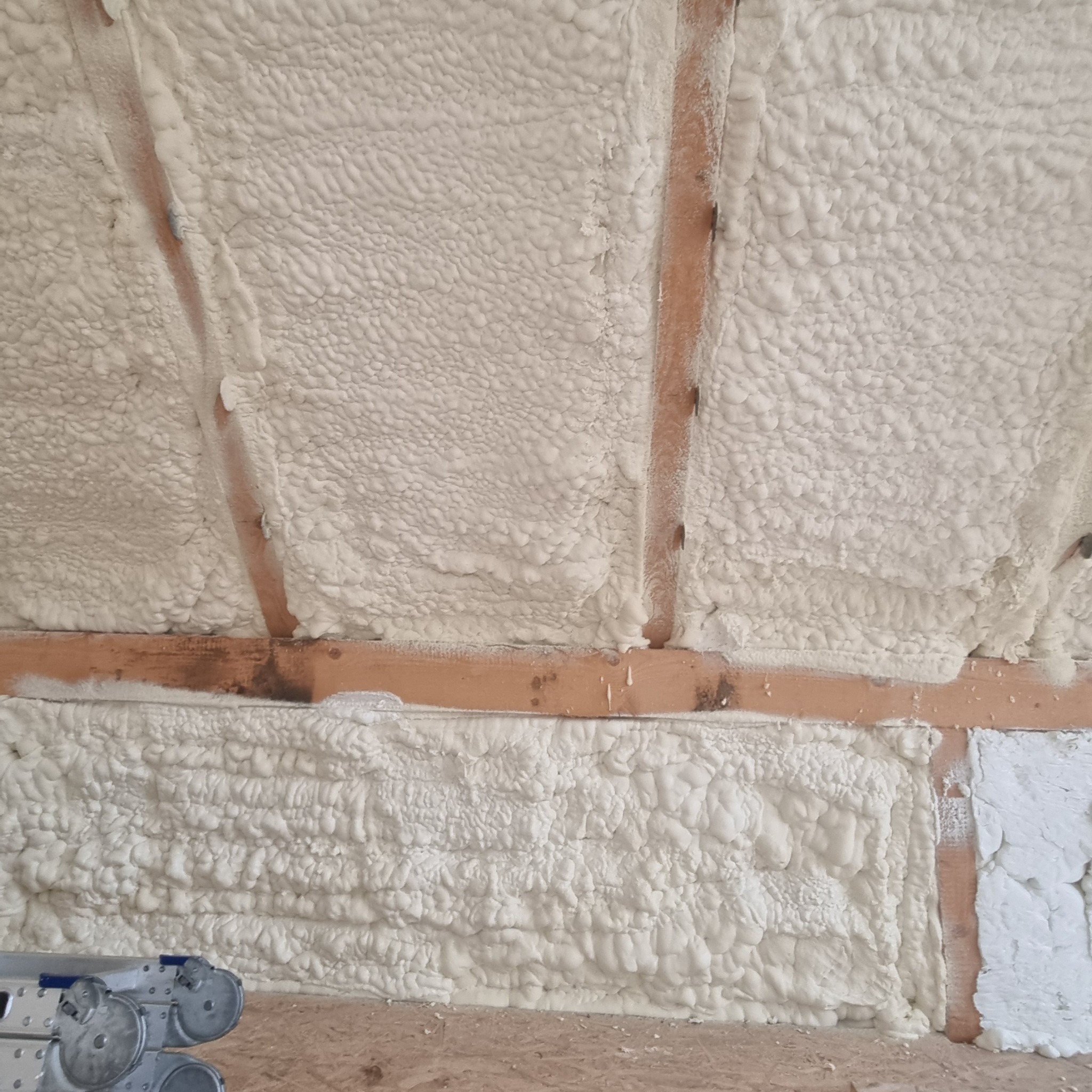 Ocieplenie poddasza pianką PUR, widoczne drewniane belki i struktura pianki. Na dole fragment drabiny.