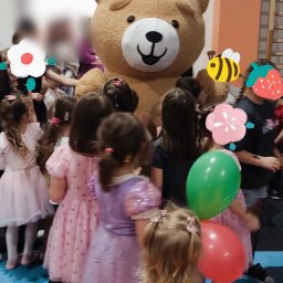 Agnieszka Bielak - Urodziny dziecka z animatorem w stroju misia. Dzieci w sukienkach otaczają pluszowego misia. Dekoracje urodzinowe i kolorowe balony. Sala z niebieską podłogą.