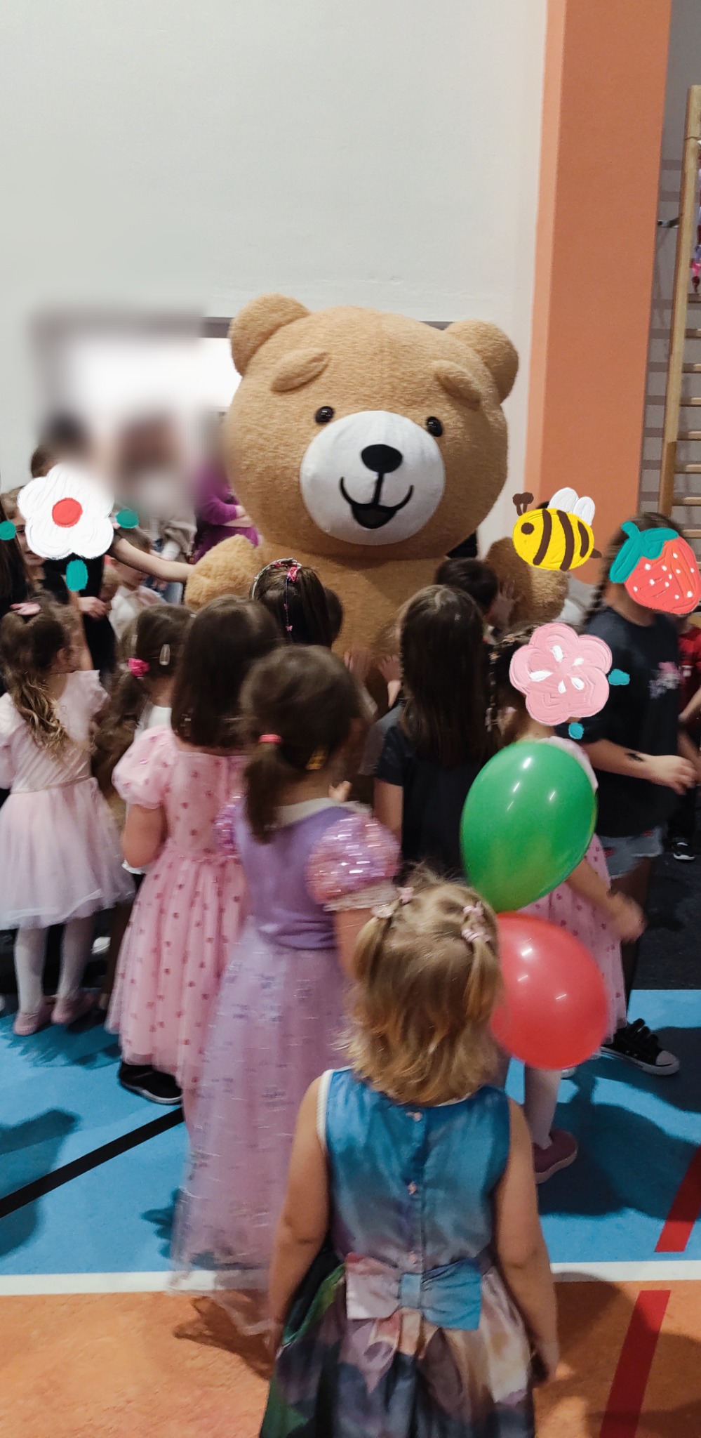 Urodziny dziecka z animatorem w stroju misia. Dzieci w sukienkach otaczają pluszowego misia. Dekoracje urodzinowe i kolorowe balony. Sala z niebieską podłogą.