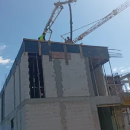 Budowa domu z betonu komórkowego: pracownicy obsługują pompę do betonu, w tle żuraw budowlany i błękitne niebo. Widok z dołu na dwukondygnacyjny budynek.