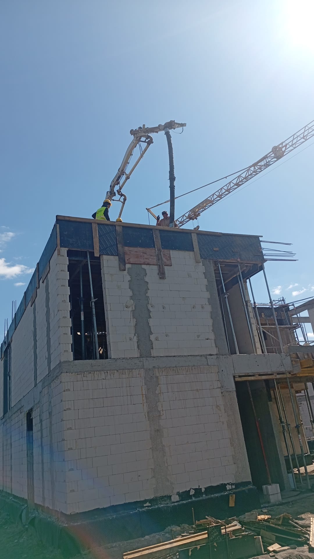 Budowa domu z betonu komórkowego: pracownicy obsługują pompę do betonu, w tle żuraw budowlany i błękitne niebo. Widok z dołu na dwukondygnacyjny budynek.