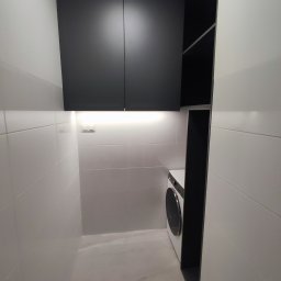PB DOMY SPÓŁKA Z OGRANICZONĄ ODPOWIEDZIALNOŚCIĄ - Nowoczesna pralnia z czarnymi szafkami na wymiar i pralką, oświetlona listwą LED. Minimalistyczny design, białe płytki na ścianach i podłodze. Funkcjonalne rozwiązanie.