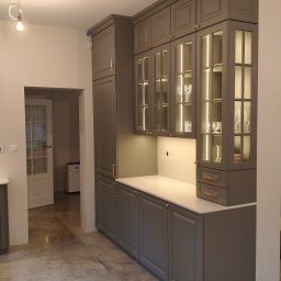 PB DOMY SPÓŁKA Z OGRANICZONĄ ODPOWIEDZIALNOŚCIĄ - Szare meble kuchenne z przeszklonymi szafkami i oświetleniem LED w Czarnocinie. Klasyczny styl, złote uchwyty, biały blat. Widok na otwarte drzwi.