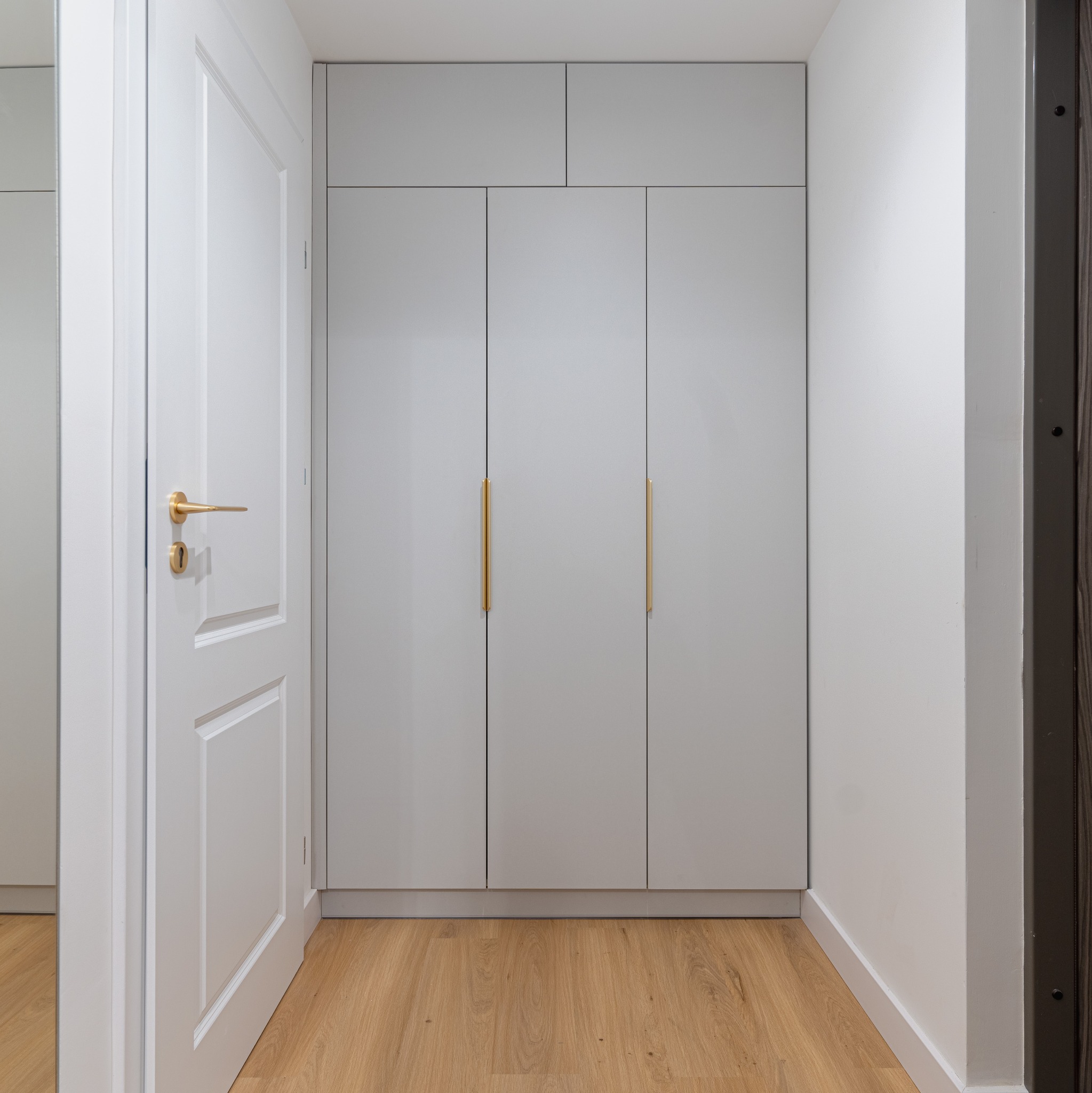 Jasnoszara szafa wnękowa z trzema drzwiami i złotymi uchwytami, widoczna z perspektywy wnętrza, z fragmentem białych drzwi po lewej stronie i drewnianą podłogą.