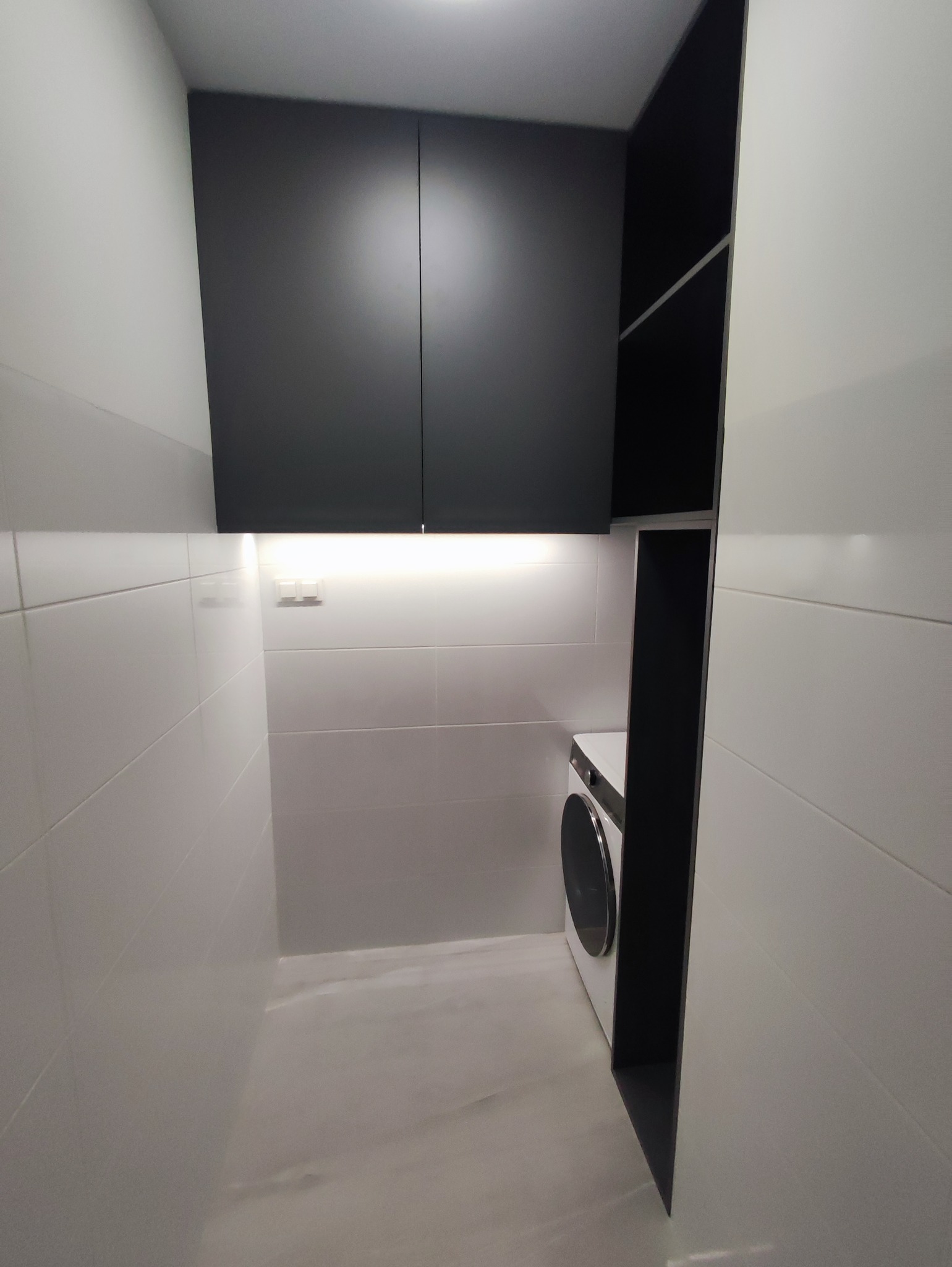 Nowoczesna pralnia z czarnymi szafkami na wymiar i pralką, oświetlona listwą LED. Minimalistyczny design, białe płytki na ścianach i podłodze. Funkcjonalne rozwiązanie.