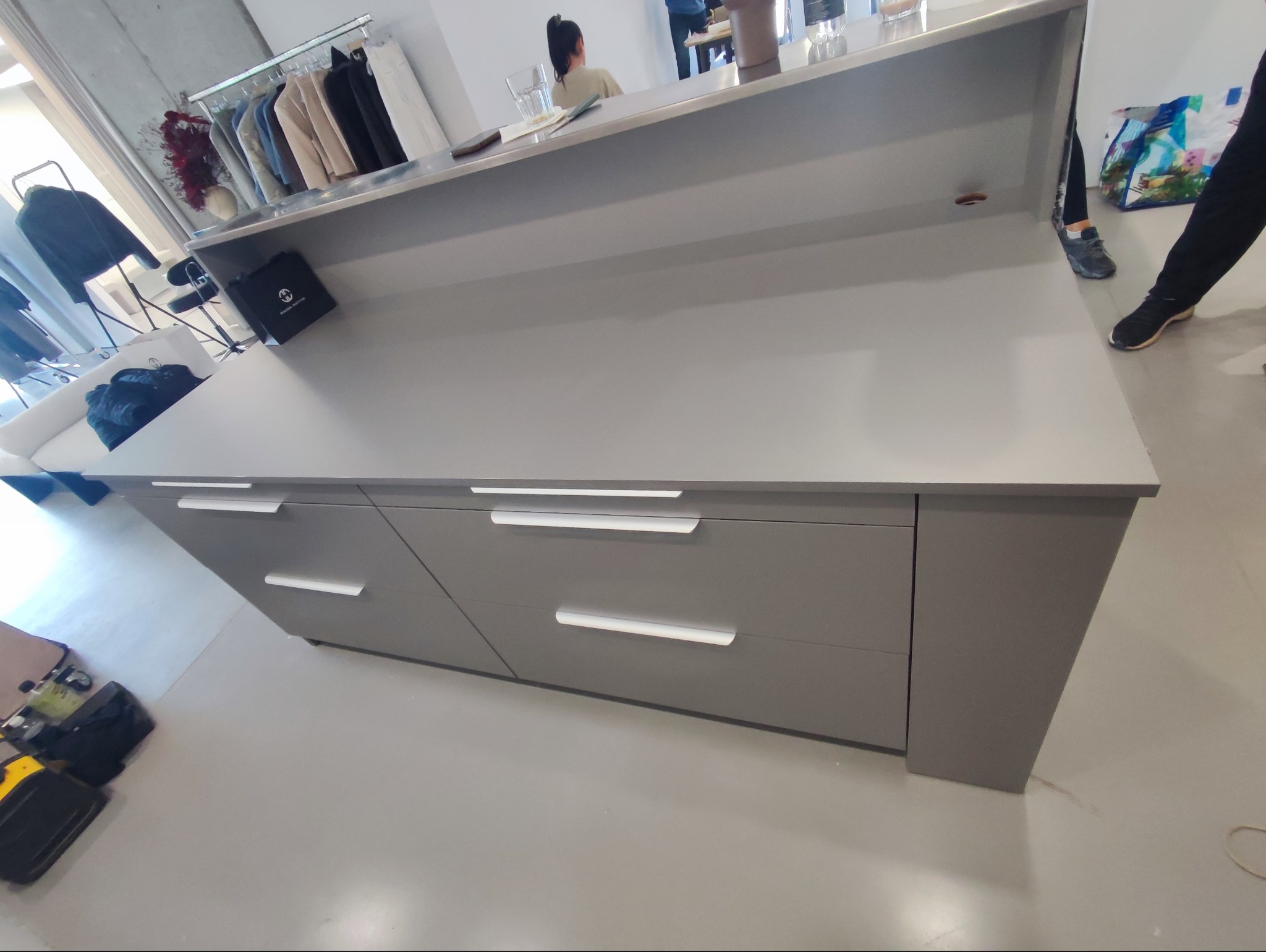 Szara lada recepcyjna z trzema szufladami i białymi uchwytami. W tle wieszak z ubraniami i osoba. Widoczne narzędzia na podłodze.