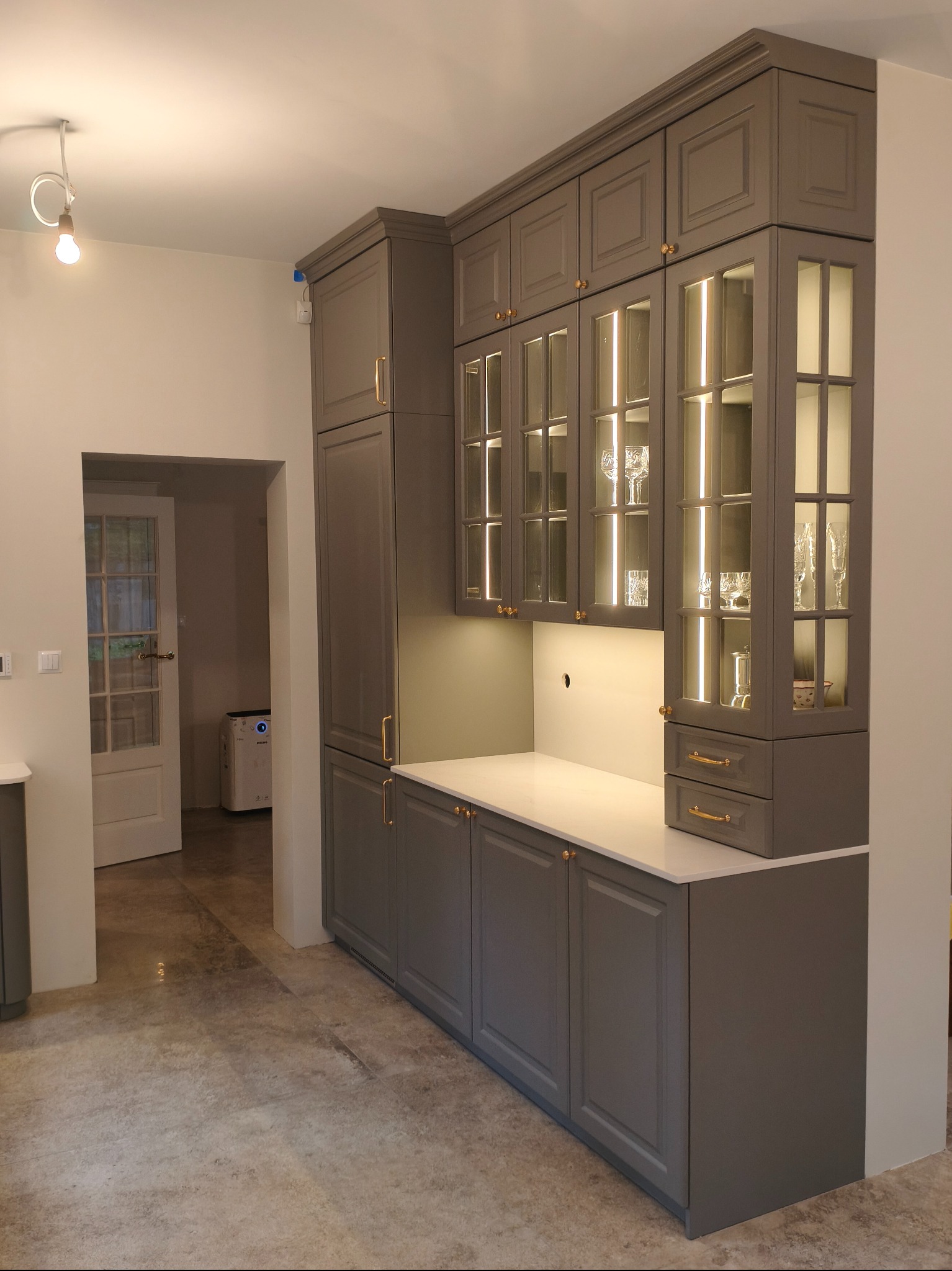 Szare meble kuchenne z przeszklonymi szafkami i oświetleniem LED w Czarnocinie. Klasyczny styl, złote uchwyty, biały blat. Widok na otwarte drzwi.