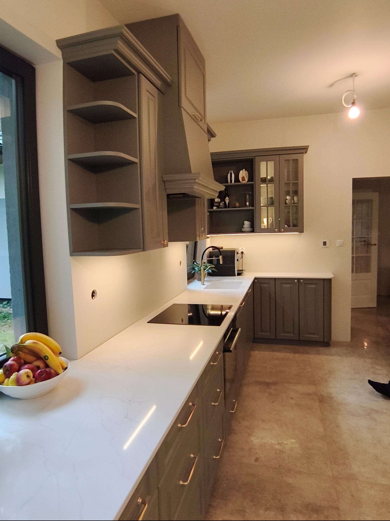 Szara kuchnia z białym blatem i owocami w misce przy oknie. Górne szafki z półkami i przeszkleniami. Nowoczesny design z oświetleniem podszafkowym.