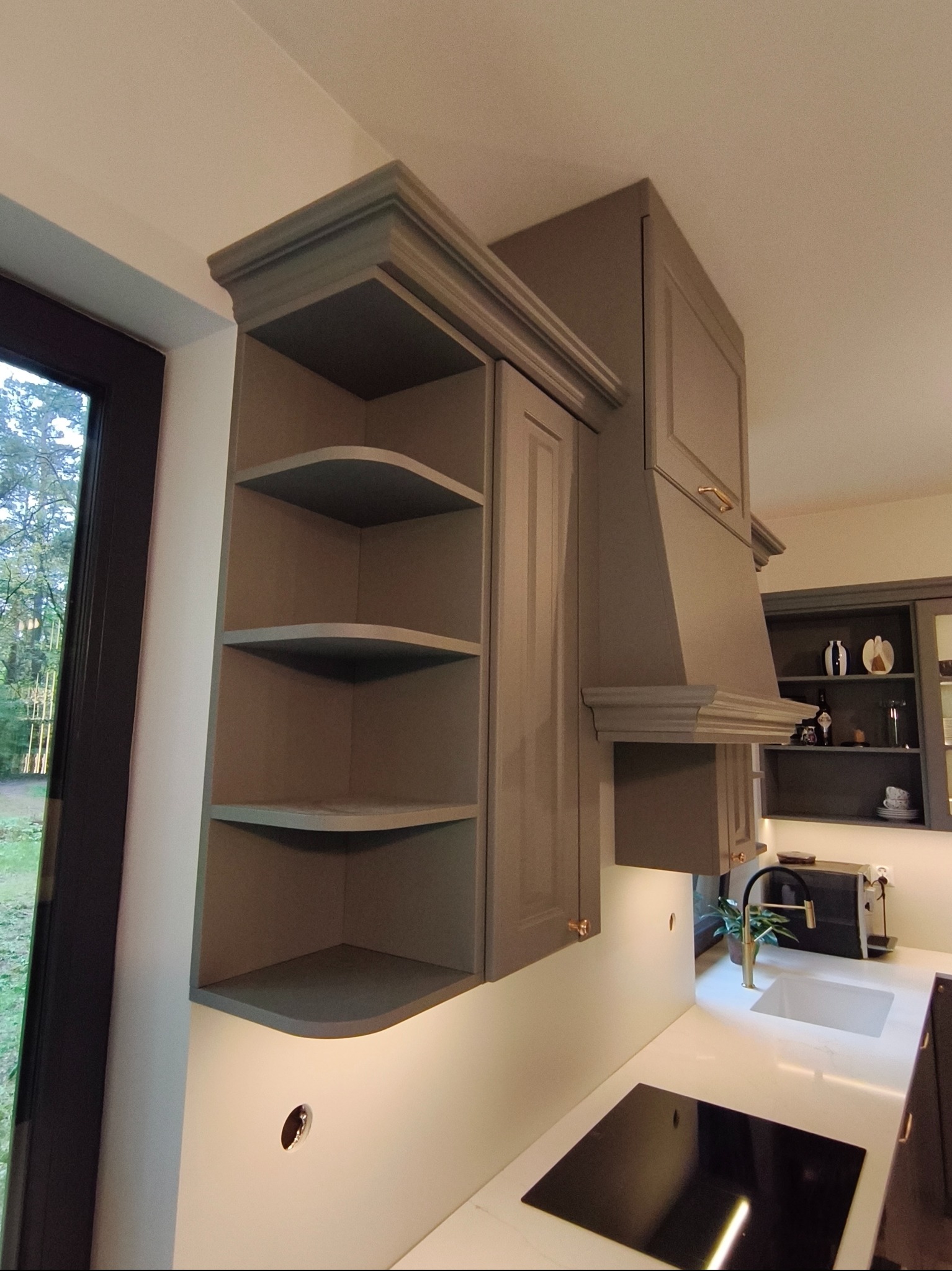 Szara zabudowa kuchenna z otwartymi półkami narożnymi i okapem, widok z dołu, z blatem i płytą indukcyjną. W tle szafka z dekoracjami.