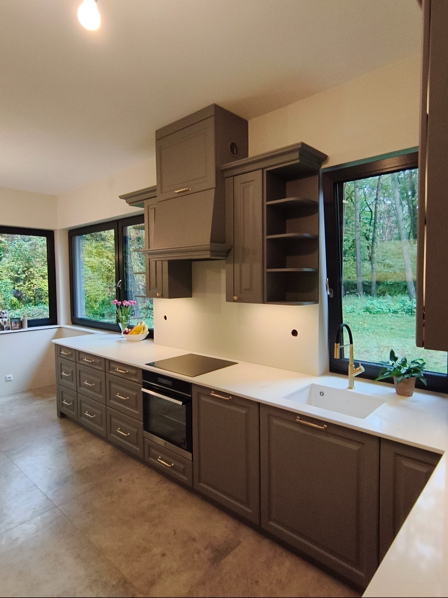 Szara kuchnia na wymiar z frezowanymi frontami, złotymi uchwytami i blatem z konglomeratu, z widokiem na zieleń za oknem. Nowoczesny design z elementami klasycznymi.