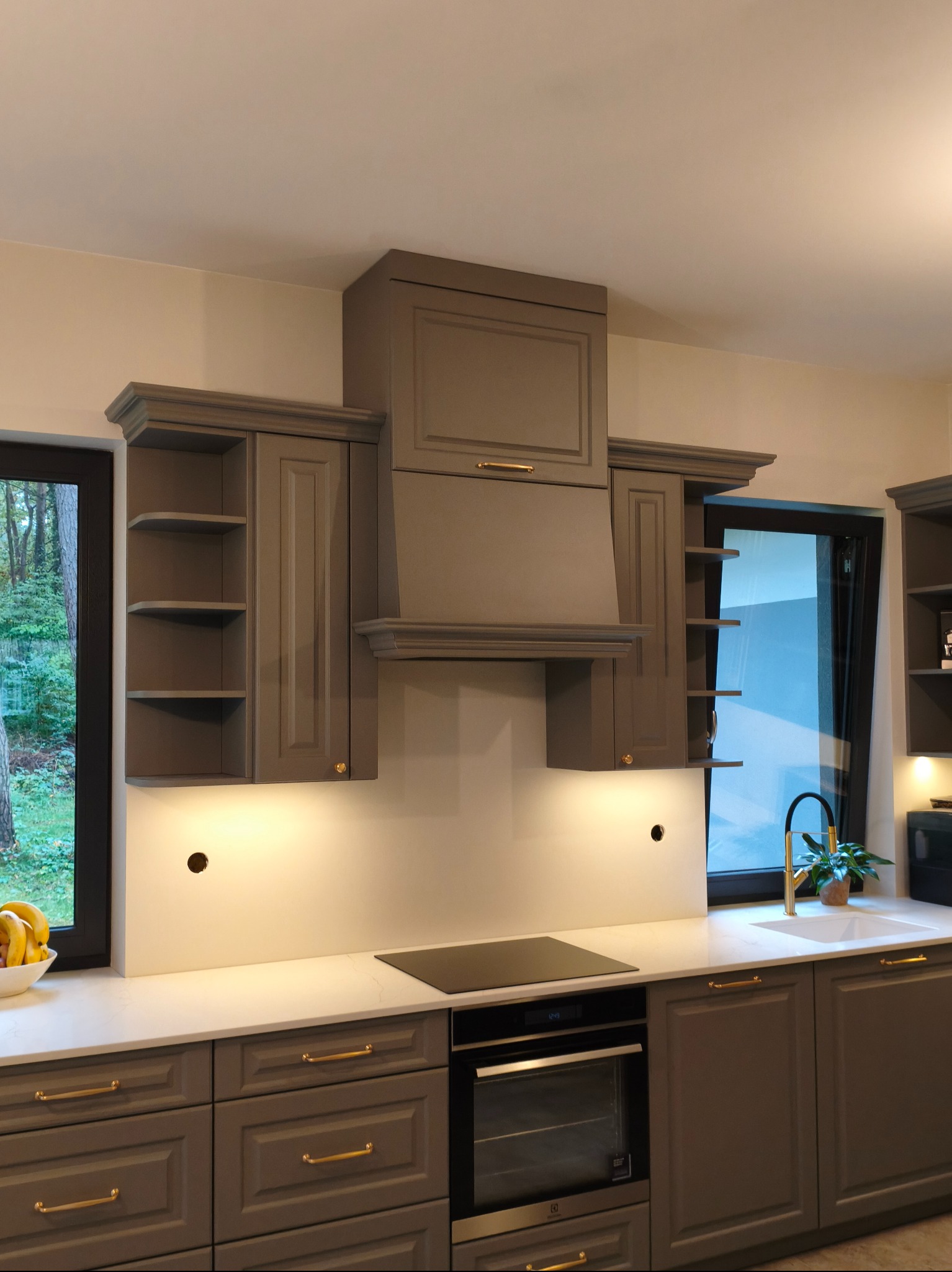 Szara kuchnia z frezowanymi frontami, złotymi uchwytami i białym blatem. Nad płytą grzewczą zabudowany okap. Po bokach okapu szafki z półkami. Widok na zieleń za oknem.