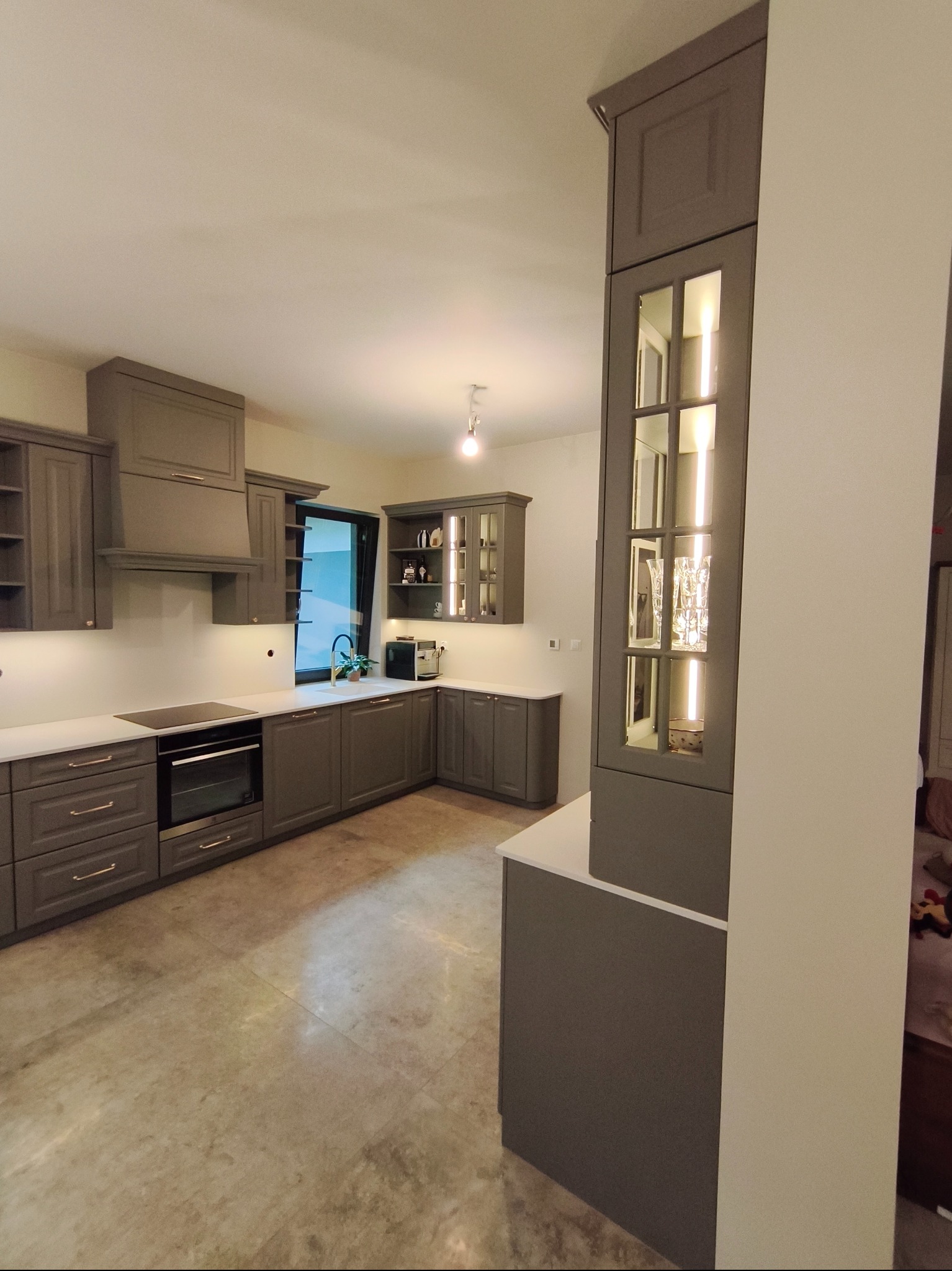 Szara kuchnia z oświetleniem LED w szklanych witrynach, biały blat, nowoczesny design, widok na okno i sprzęt AGD. Uwagę przykuwa wysoka zabudowa z przeszkleniami.