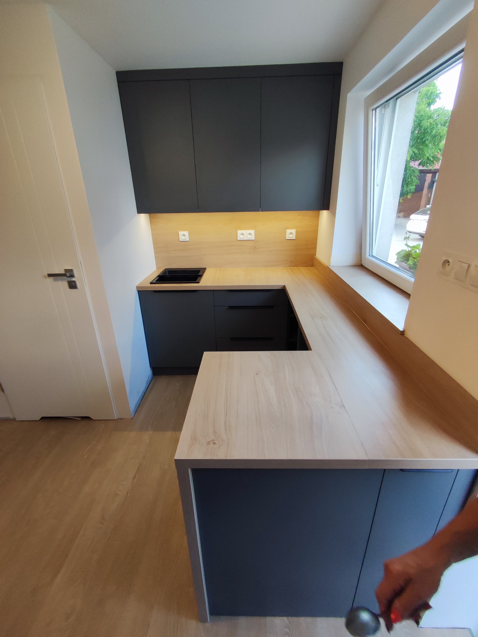 Nowoczesna kuchnia w kształcie litery U z szarymi szafkami i drewnianym blatem, oświetlona od spodu, z oknem w tle. Widoczny fragment osoby.