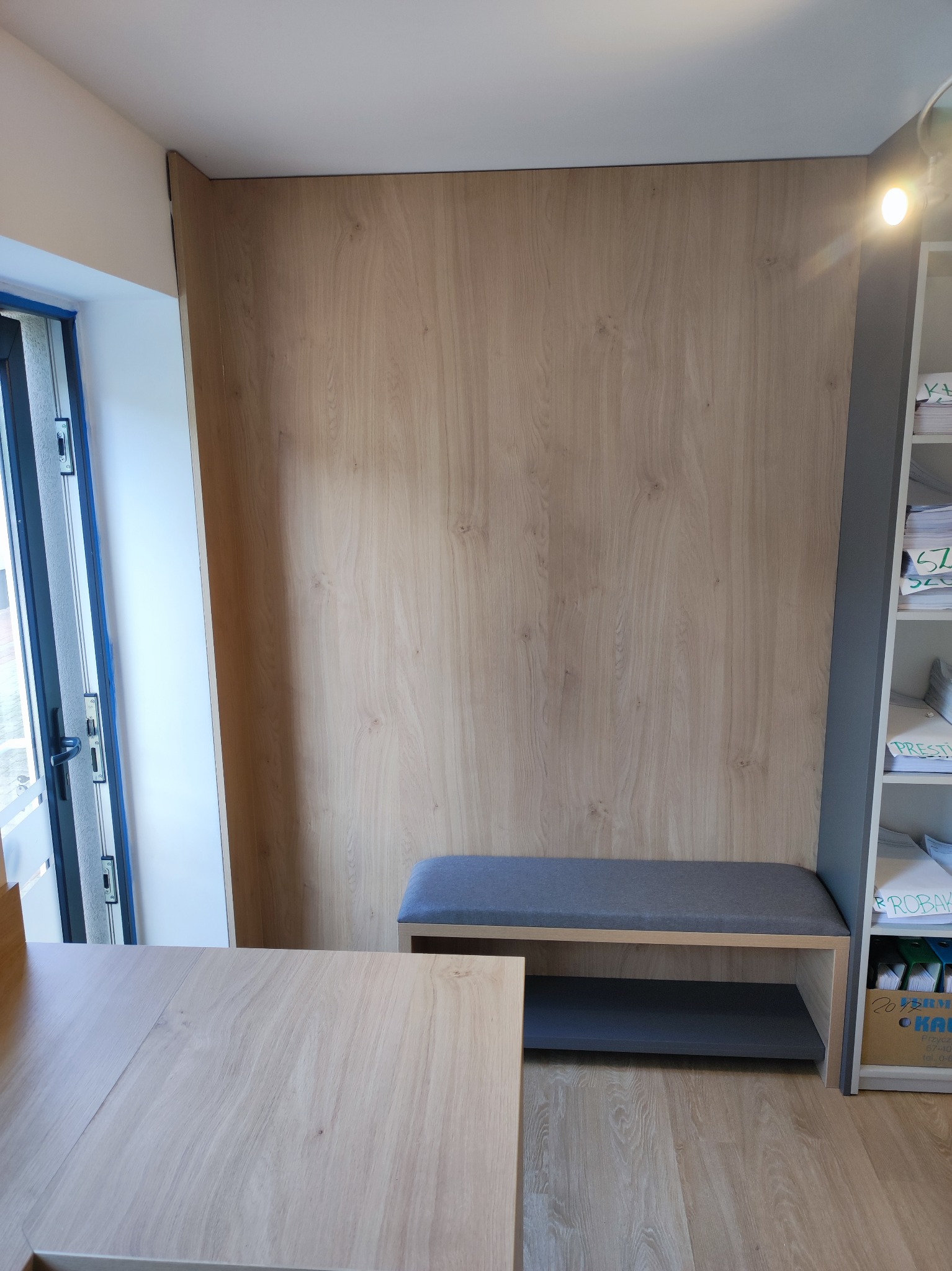 Drewniana zabudowa wnęki z siedziskiem i półką, dopasowana kolorystycznie do podłogi i biurka. Obok regał z dokumentami. Widok z perspektywy osoby stojącej.
