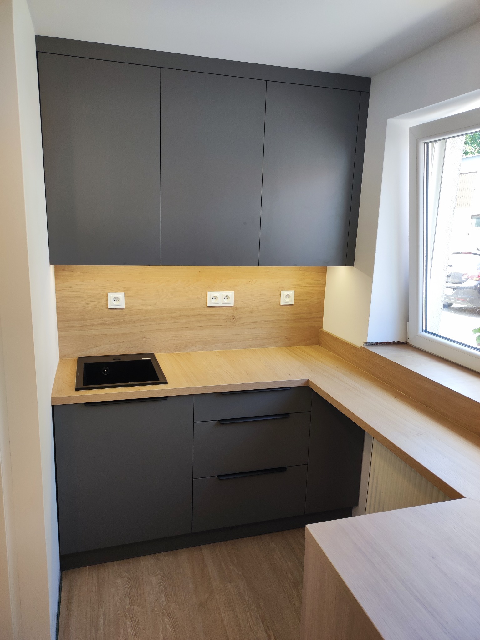 Nowoczesna kuchnia z szarymi szafkami i drewnianym blatem, oświetlona punktowo, z czarnym zlewem i gniazdkami elektrycznymi na ścianie. Widok na okno z zewnątrz.