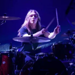Perkusista z długimi włosami w trakcie występu, uderza w talerze. Widoczne bębny i pałeczki. Scena oświetlona niebieskim światłem.
