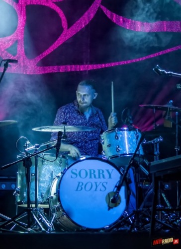 Perkusista w akcji na scenie, bębny z napisem 'SORRY BOYS', intensywne oświetlenie sceniczne, dym, mikrofon. Artysta skupiony na grze, dynamiczne ujęcie.